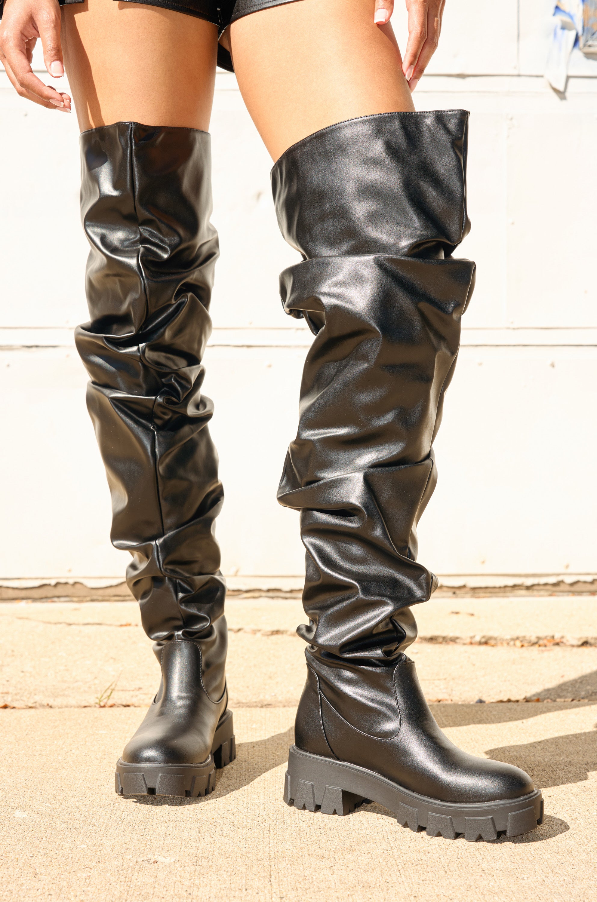 A person wears the AZALEA WANG KIPLING BLACK RUCHED UP FLAT BOOT—black faux leather, thigh-high, flat lug sole boots—scrunched and styled with black shorts for a bold, modern look on a concrete background.