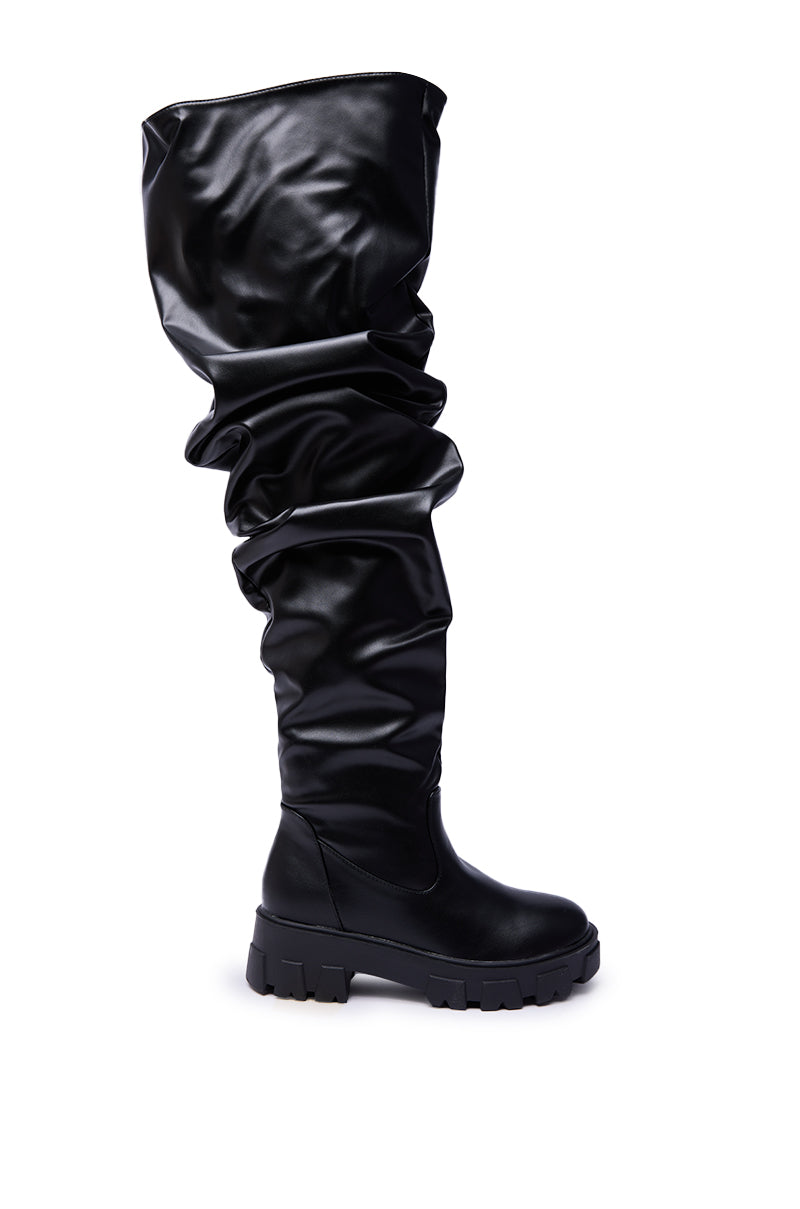 The AZALEA WANG KIPLING BLACK RUCHED UP FLAT BOOT, an over-the-knee, slouchy black faux leather style with a chunky treaded sole, is shown against a white background.