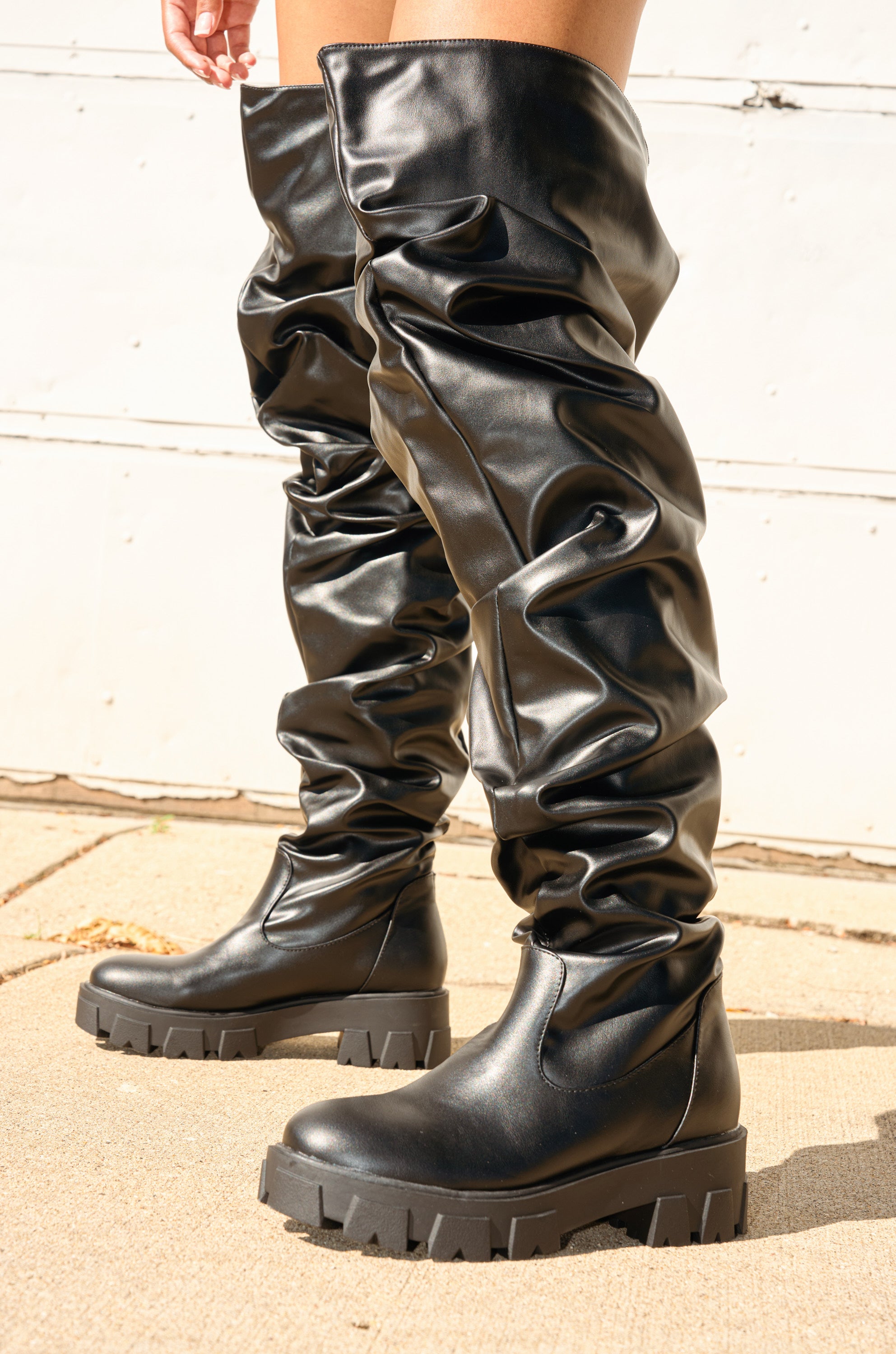Someone stands in front of a white wall on a light surface wearing the AZALEA WANG KIPLING Black Ruched Up Flat Boot—shiny, black, faux leather knee-high boots featuring thick, chunky soles and ruched detailing.