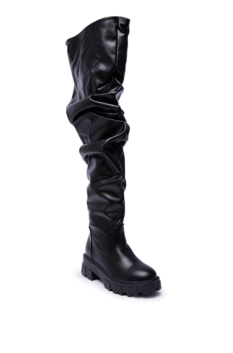 The AZALEA WANG KIPLING BLACK RUCHED UP FLAT BOOT is a shiny black faux leather thigh-high with a chunky sole and slouchy, ruched detailing, shown against a white background.