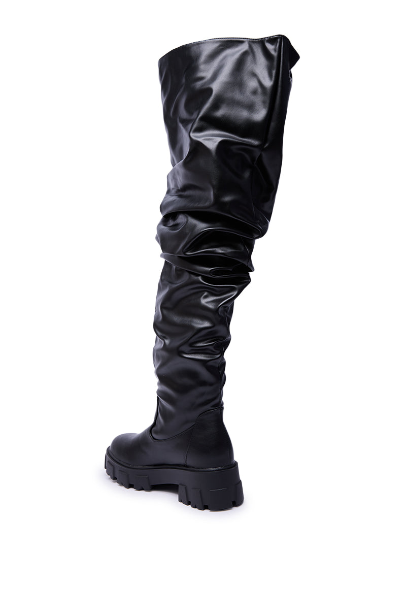 The AZALEA WANG KIPLING BLACK RUCHED UP FLAT BOOT, a single knee-high black boot in shiny faux leather with ruched detailing and a chunky sole, is displayed against a white background.