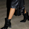 A person wearing a black leather skirt and sleek AZALEA WANG KIRIGAN black patent booties struts on a concrete floor, holding a black handbag with a silver chain and clasp.