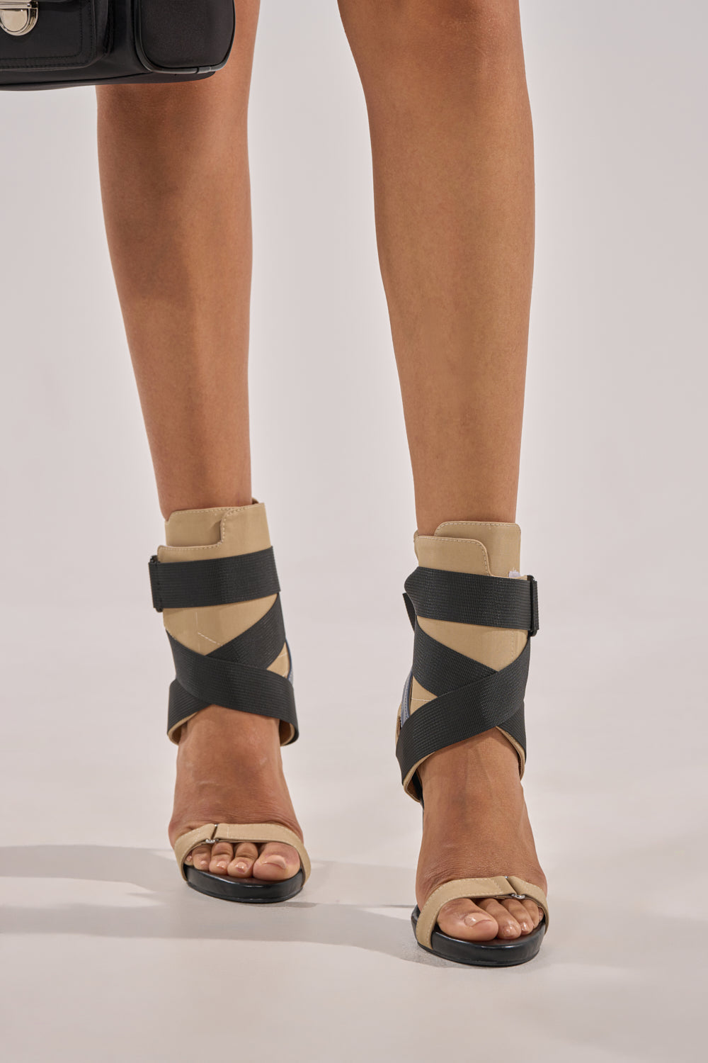 A person models the AZALEA WANG KIVA KHAKI STILETTO SPORTY SANDAL with wide black crisscross elastic straps and a comfort cushion insole, standing on a light background. A hint of a black bag appears in the corner. 2