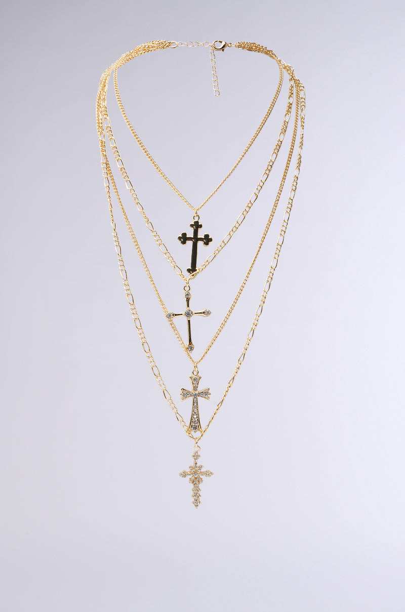 The CROSS LAYERED NECKLACE features four pendants on unique faux gold chains, including a rhinestone-studded cross, elegantly displayed on a plain gray background. 0