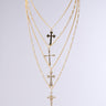 The CROSS LAYERED NECKLACE features four pendants on unique faux gold chains, including a rhinestone-studded cross, elegantly displayed on a plain gray background.
