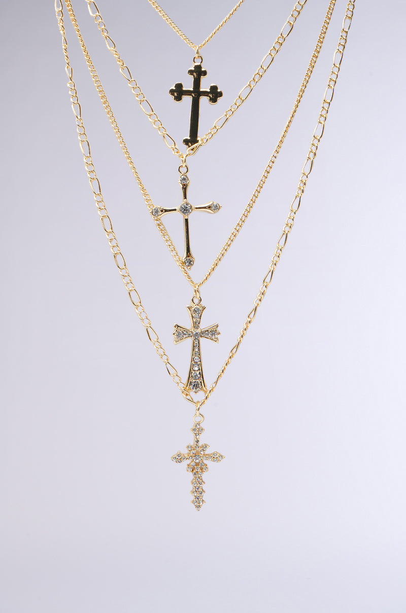 A collection of CROSS LAYERED NECKLACES hangs against a plain background, featuring faux gold crosses with rhinestones. Each cross varies in design and size, showcasing intricate details for a layered and elegant look. 2