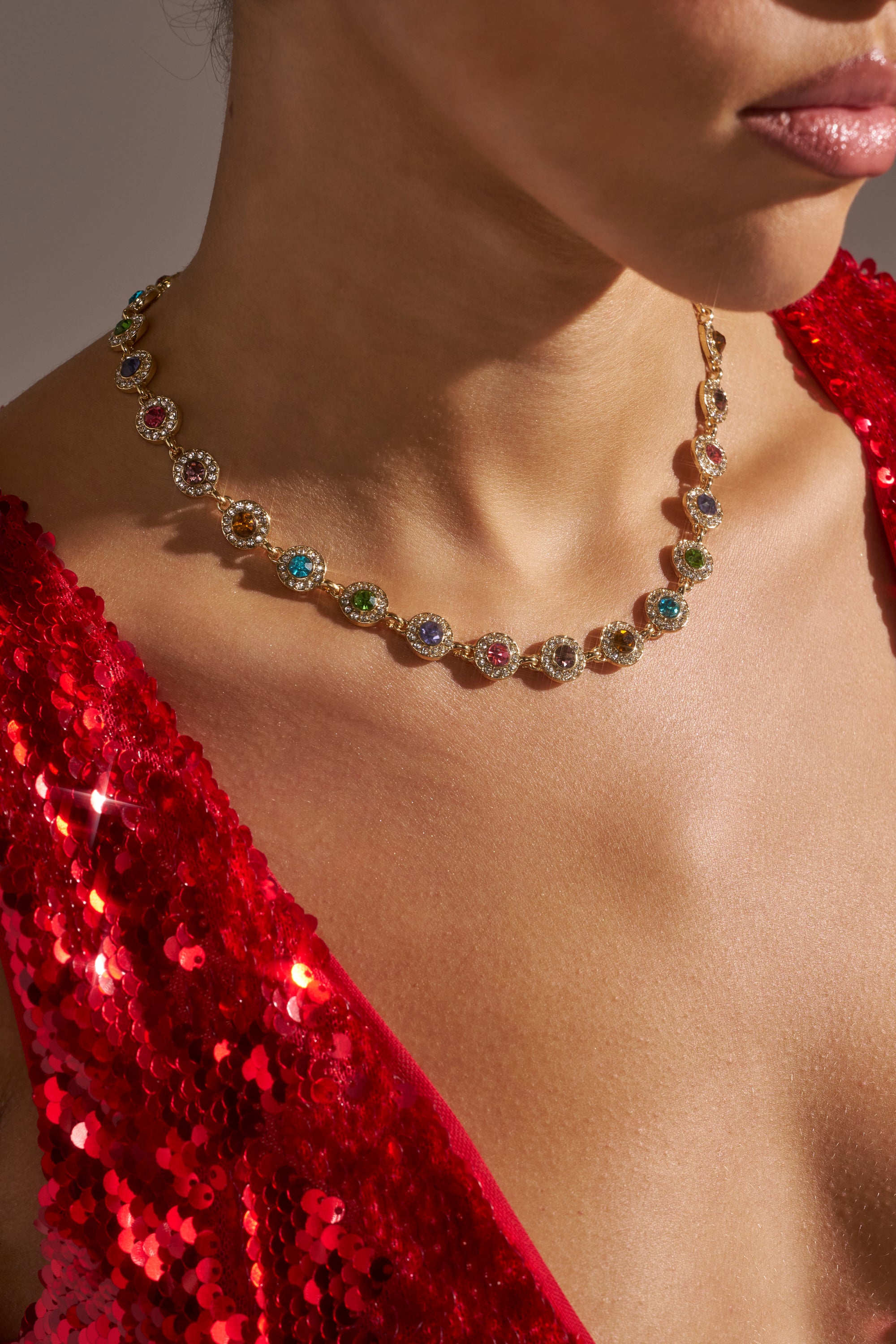 A woman wears the ITS GIVING LUXURY CHOKER, a luxury embellished necklace with multicolored gemstones, paired with a sparkling red sequin dress. The image is cropped to highlight her neck, collarbone, lips, and chest. 1