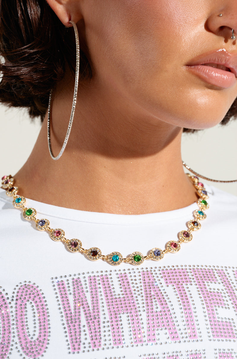 Close-up of a woman wearing the ITS GIVING LUXURY CHOKER, featuring a gold chain with multicolor gems, paired with large hoop earrings. She is also wearing a white shirt embellished with rhinestone trim and partially visible text. 3