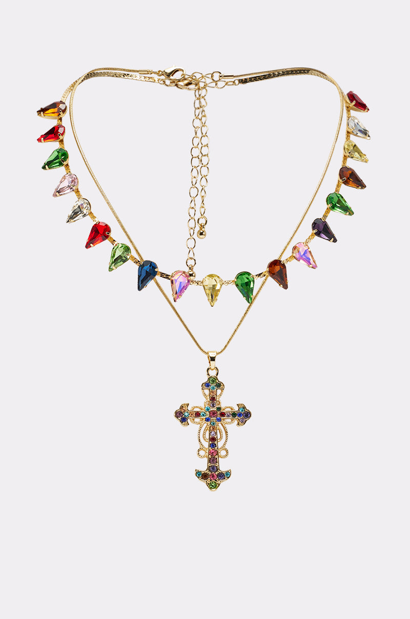 The DOLCE VITA NECKLACE is a layered piece with faceted gemstone charms and a vibrant gemstone cross pendant, adorned with multicolored gems, showcased on a simple light background. 0