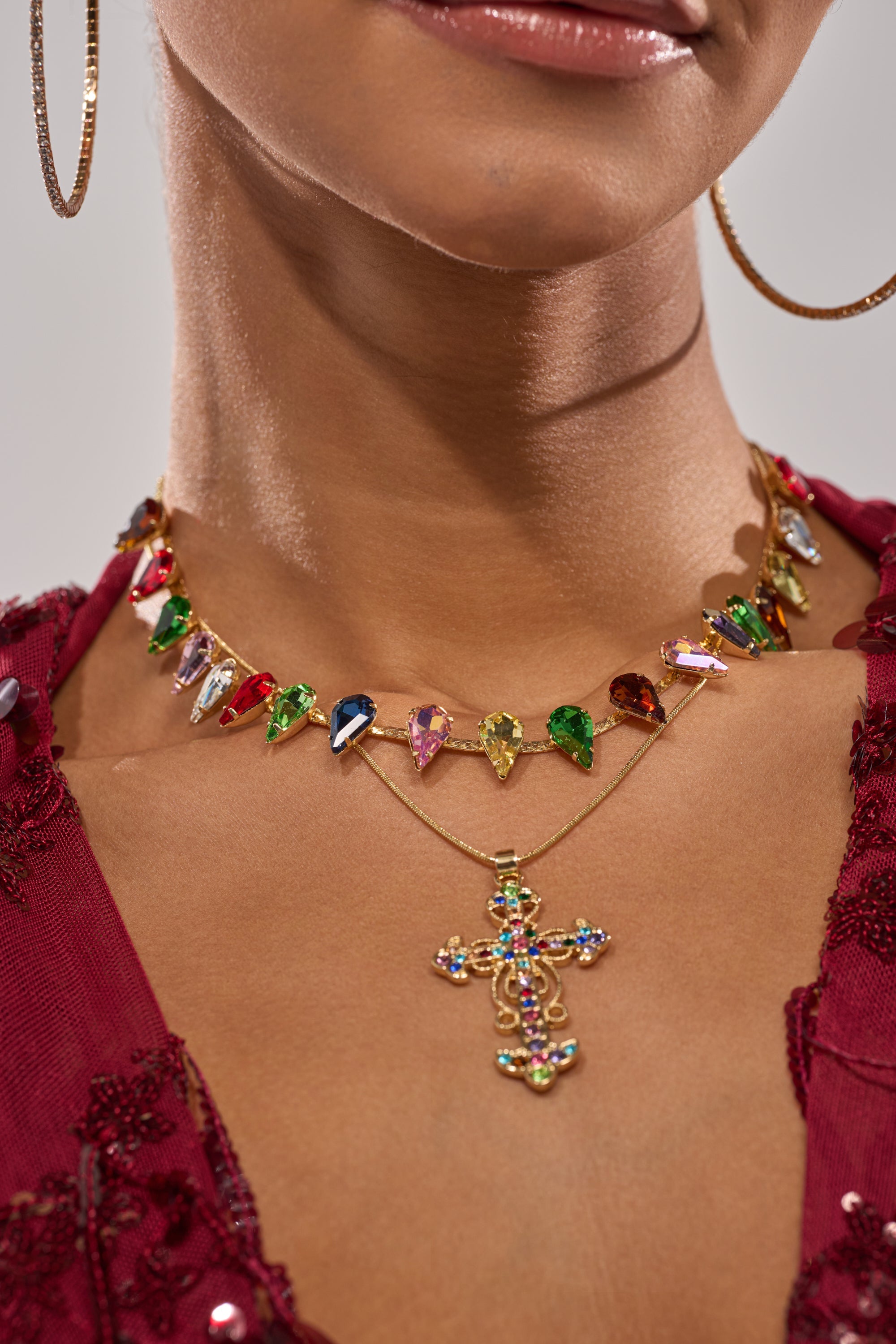 A woman wears large hoop earrings, the DOLCE VITA NECKLACE with colorful gemstone charms, a layered gold necklace with a jeweled cross pendant, and a deep red embroidered top. 1