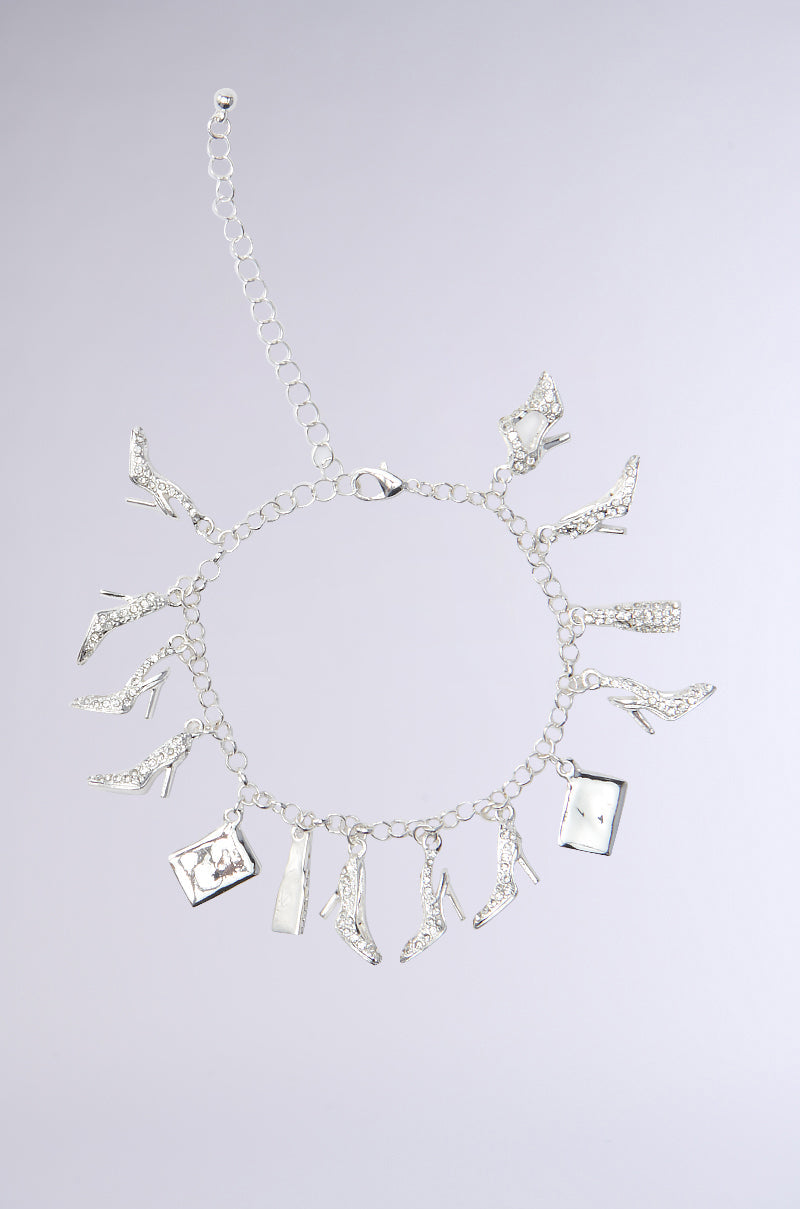 The STILETTO CHARM BRACELET features a silver-tone design with sparkling rhinestones, high-heeled shoe charms, and small rectangular accents, shown laid flat on a light gray background. 0