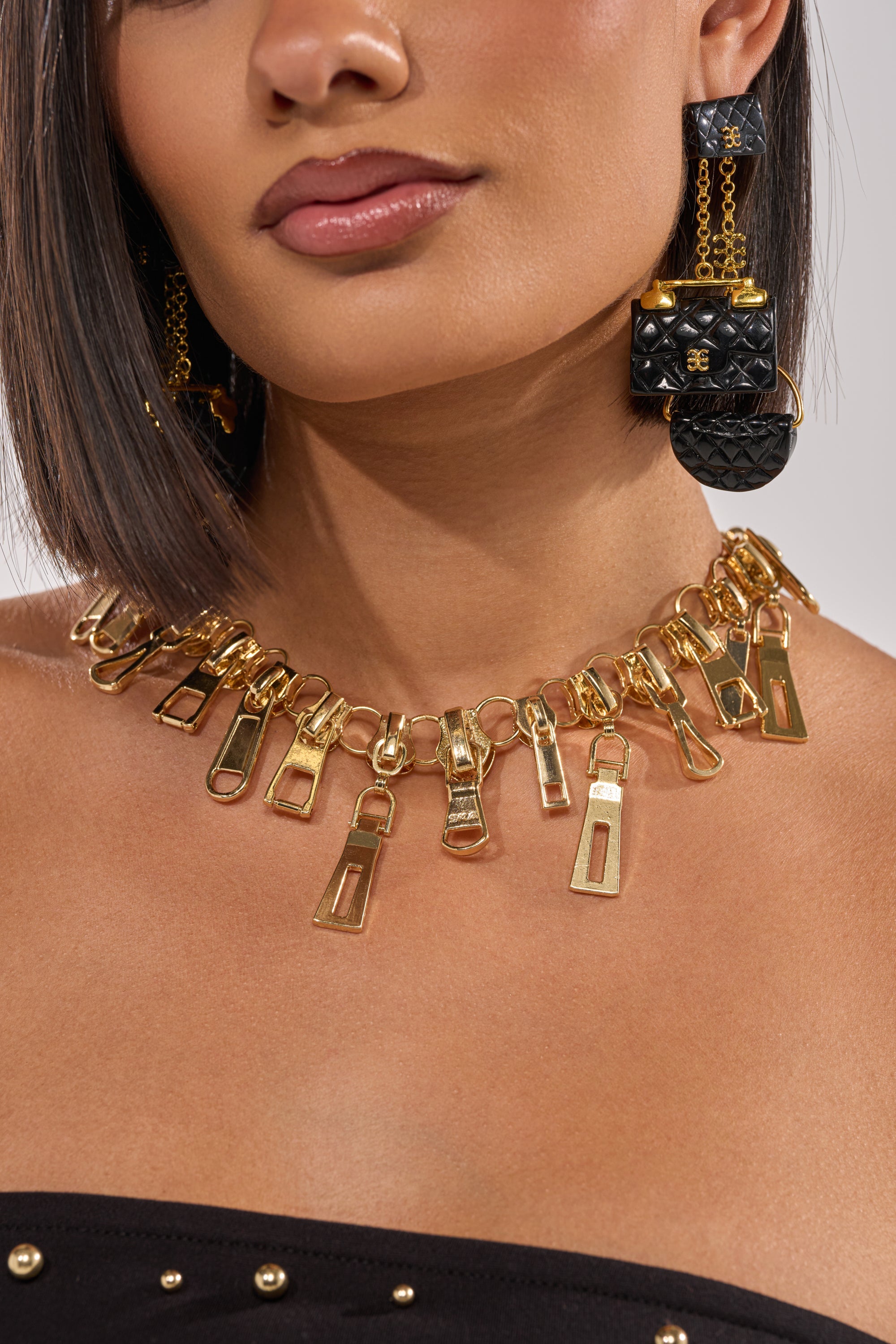 A woman in a sleek bob and a black, studded strapless top wears the ZIP IT NECKLACE, made of gold zipper pulls, and large black quilted handbag-shaped earrings. 1
