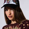 A young person with long brown hair wearing a dark red, distressed Def Leppard t-shirt poses against a plain background. They have on a black WILD THANG PATCH TRUCKER HAT featuring colorful embroidered graphics of a tiger and roses on the front panel, complete with an adjustable snapback closure.