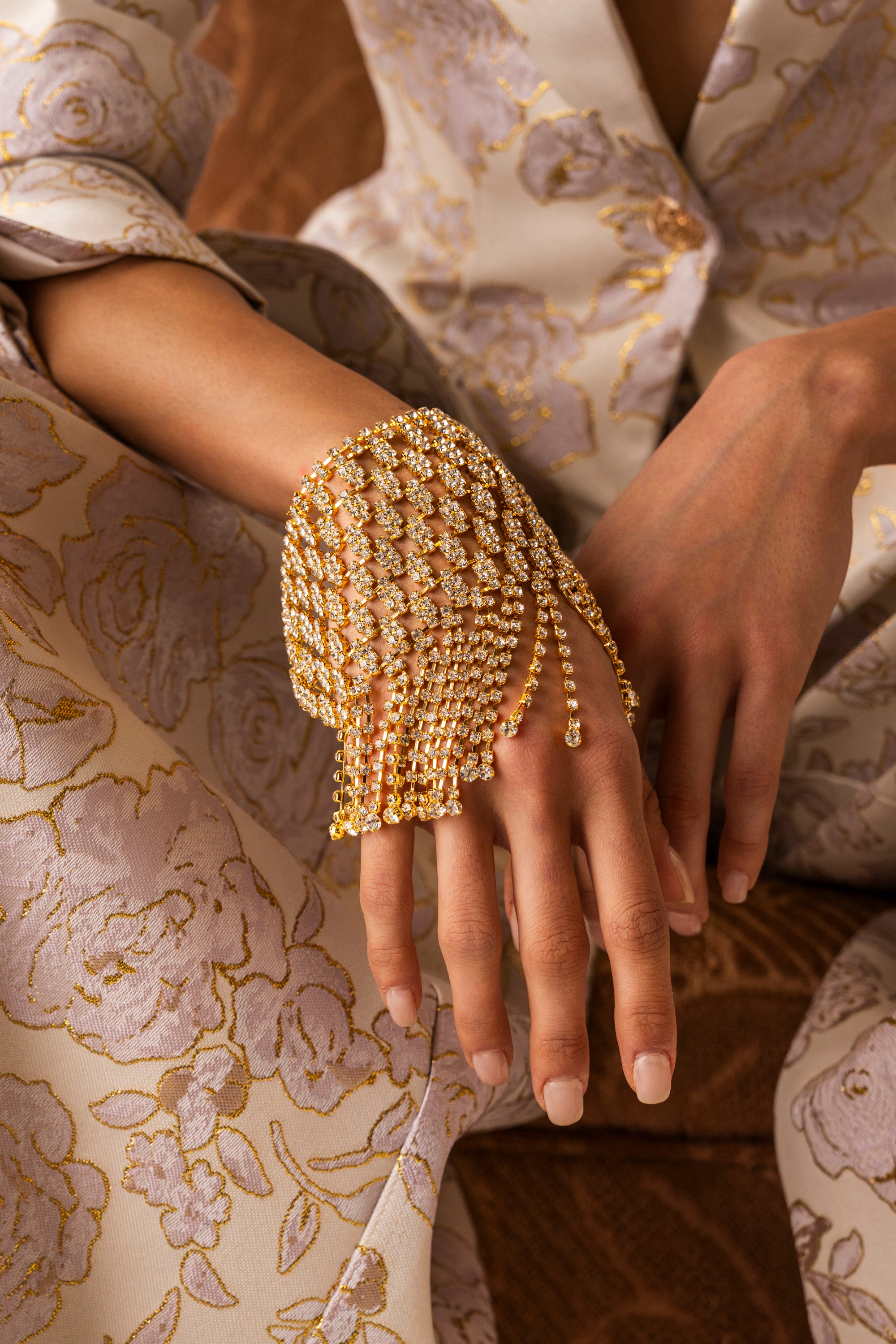 A person wears the IN MY ERA BRACELET, a gold statement piece, draped elegantly over their hand and wrist, complementing their cream and gold floral-patterned outfit. 0