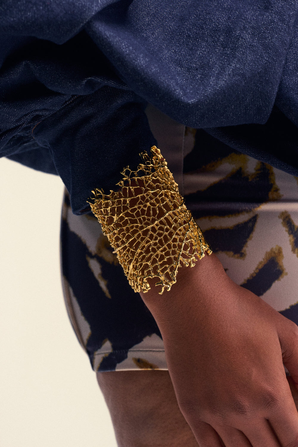 A person wearing the AT THE EVENT BRACELET CUFF—a gold, lace-patterned statement piece reminiscent of a coral reef cuff bracelet—with a dark blue sleeve and patterned shorts visible. Their hand is relaxed and slightly bent at the wrist, perfectly showcasing this summer accessory.