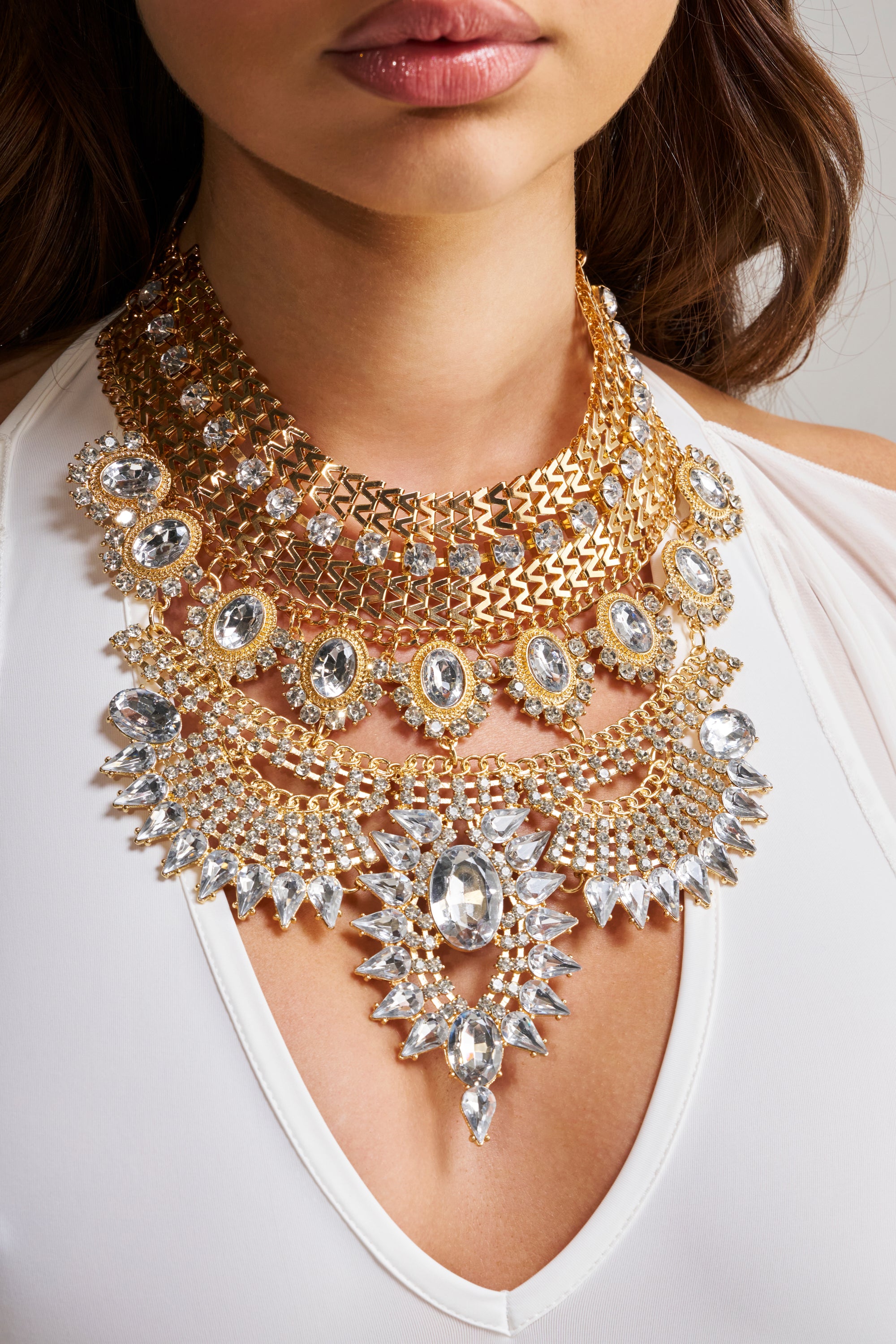 A woman in a white top with a deep neckline models the NOSTALGIA NECKLACE, a striking statement bib necklace featuring multiple layers of sparkling clear gemstones—an elegant choice for evening wear.