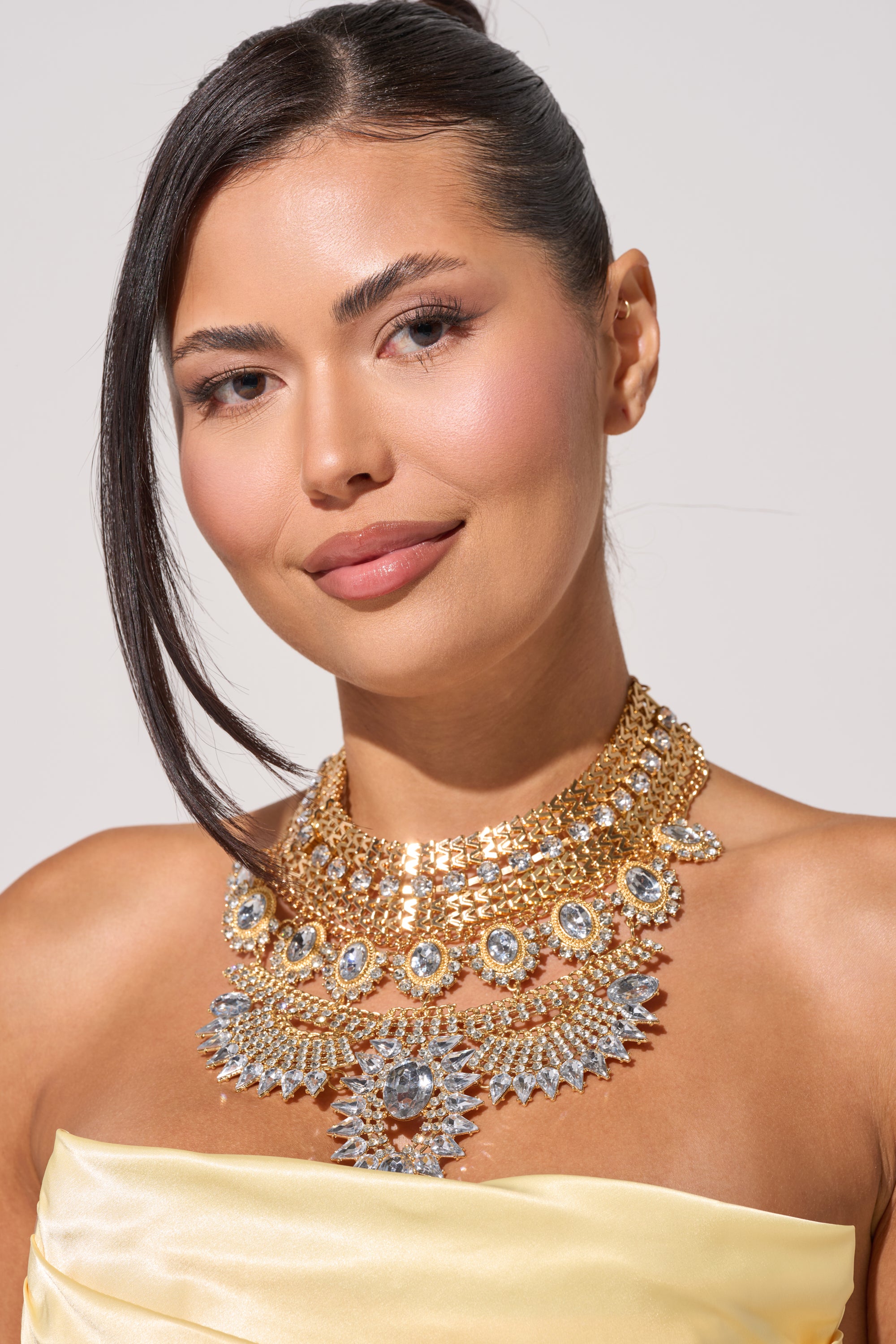 A woman with medium skin and straight dark hair in a sleek updo smiles softly, wearing a strapless yellow top and the elegant NOSTALGIA NECKLACE, featuring gold crystals, against a plain background to showcase her stunning evening jewelry. 0