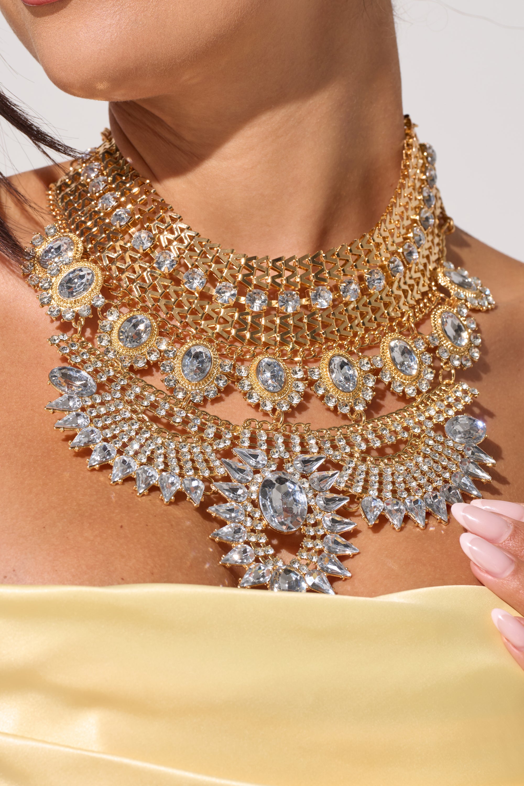 A woman in a strapless yellow dress models the NOSTALGIA NECKLACE, an elaborate statement bib adorned with large, sparkling clear gemstones and intricate detailing—perfect for glamorous evening wear. 1