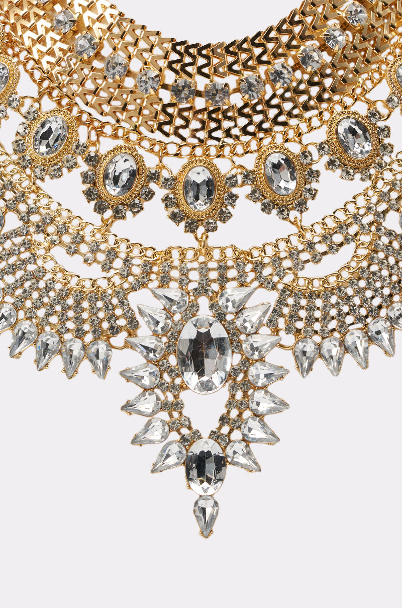 Close-up of the NOSTALGIA NECKLACE: an ornate gold statement piece with multiple rows of clear, faceted gemstones, intricate patterns, and a large central gem surrounded by smaller stones—ideal for evening wear. 3