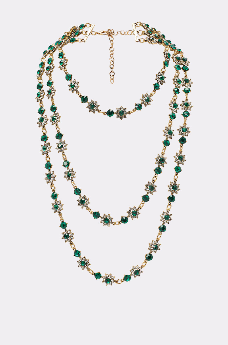 The GOOD FOR IT NECKLACE is a gold statement piece with three chains, gold-tone accents, emerald green beads, and star-shaped floral charms, showcased against a plain light background. 0