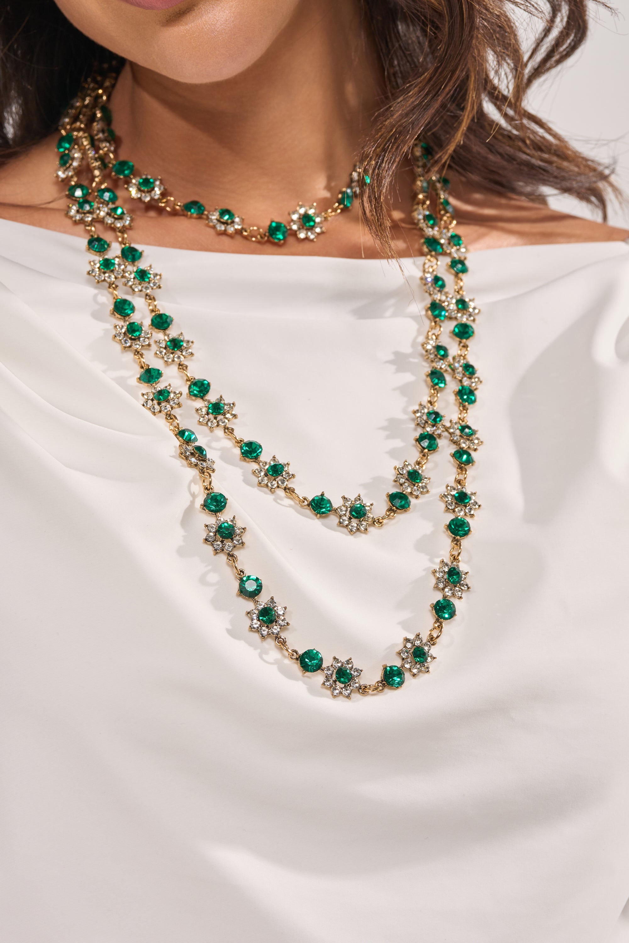 A woman wears the GOOD FOR IT NECKLACE, a gold-tone piece with emerald green gemstones and small white flower accents, elegantly layered over a white off-the-shoulder top. 1