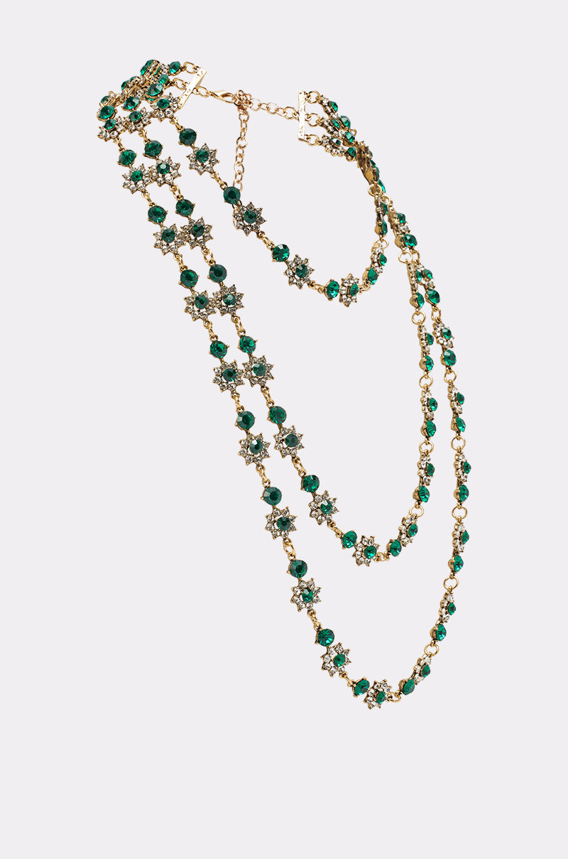 The GOOD FOR IT NECKLACE is a gold-tone layered necklace with emerald green crystal beads and small star-shaped charms, featuring an adjustable chain clasp and cascading layers. Displayed on a plain light background. 2