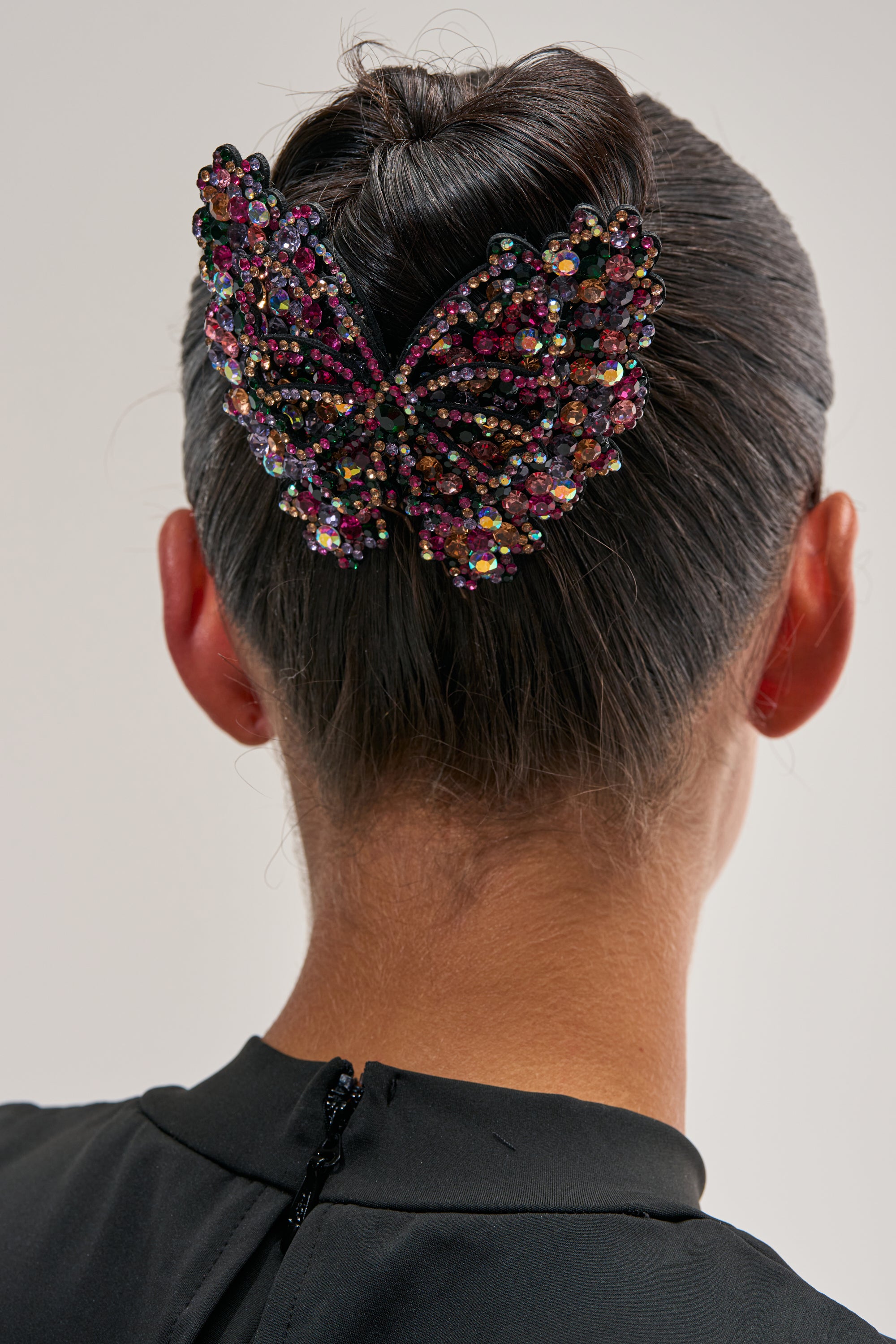 A person with dark hair styled in a bun wears the FLYING AWAY BUTTERFLY HAIR CLIP, a large and colorful rhinestone accessory. The photo is taken from behind, highlighting the sparkling clip as well as the back of their head and neck.