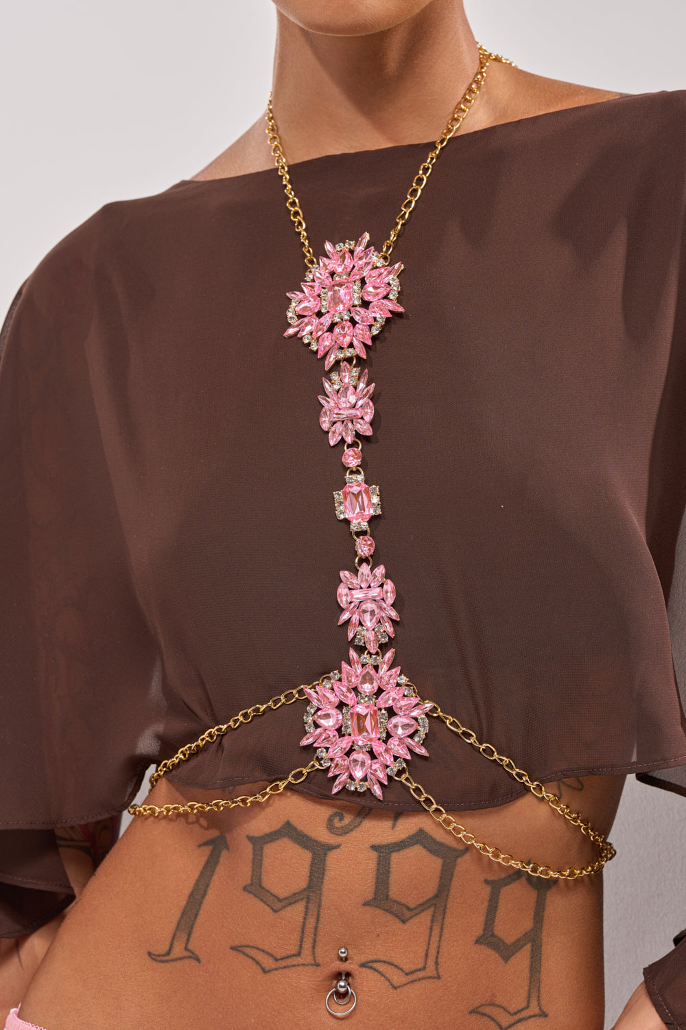 A person wearing a brown cropped top and the PRETTY IN PINK BODY CHAIN with gold festival jewelry draped across their chest and waist. A "1999" tattoo and navel piercing add to the luxe layered look. 0