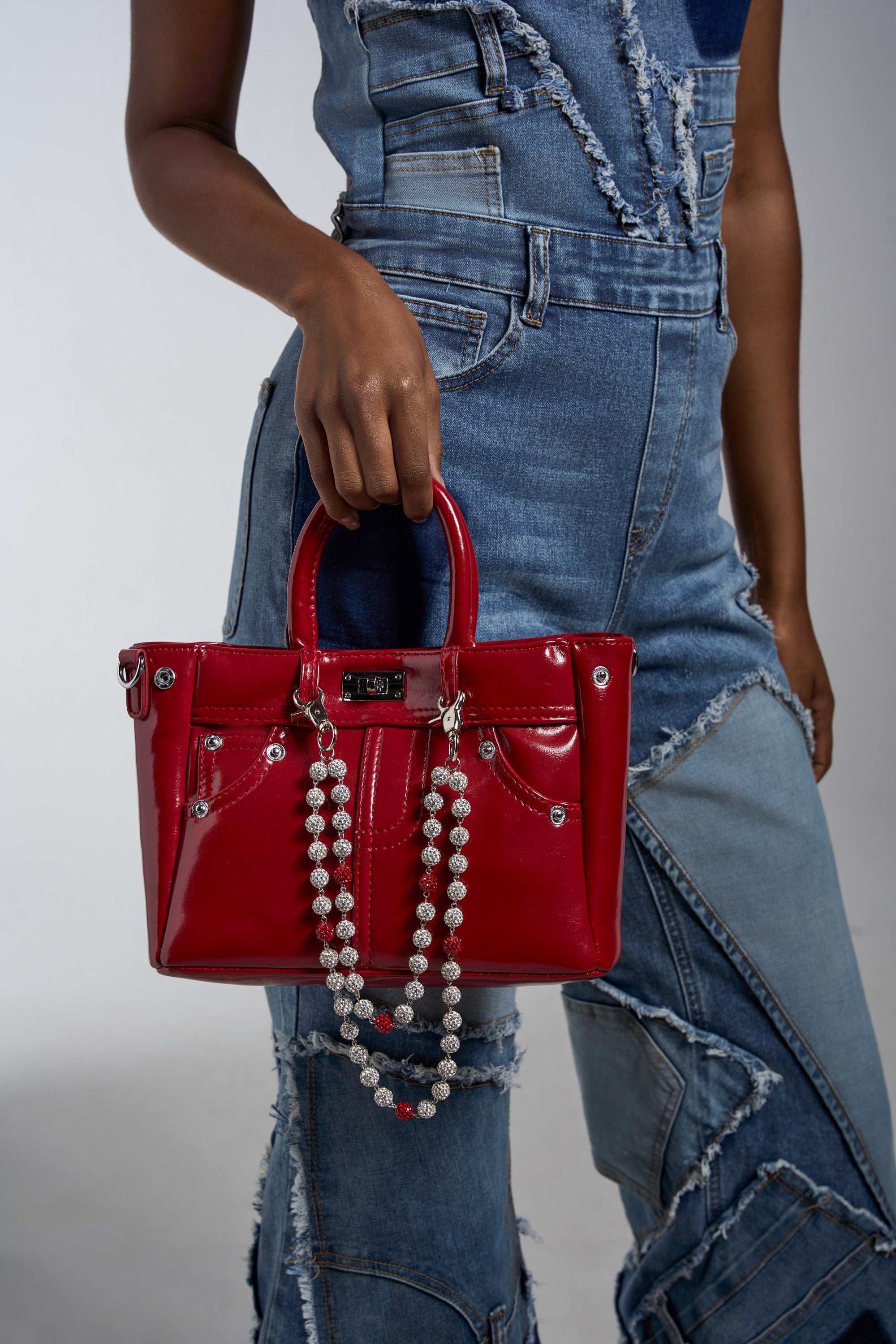 A person holds a shiny red handbag adorned with the BLING PANT & BAG CHAIN rhinestone accessory, standing against a plain light background. 0