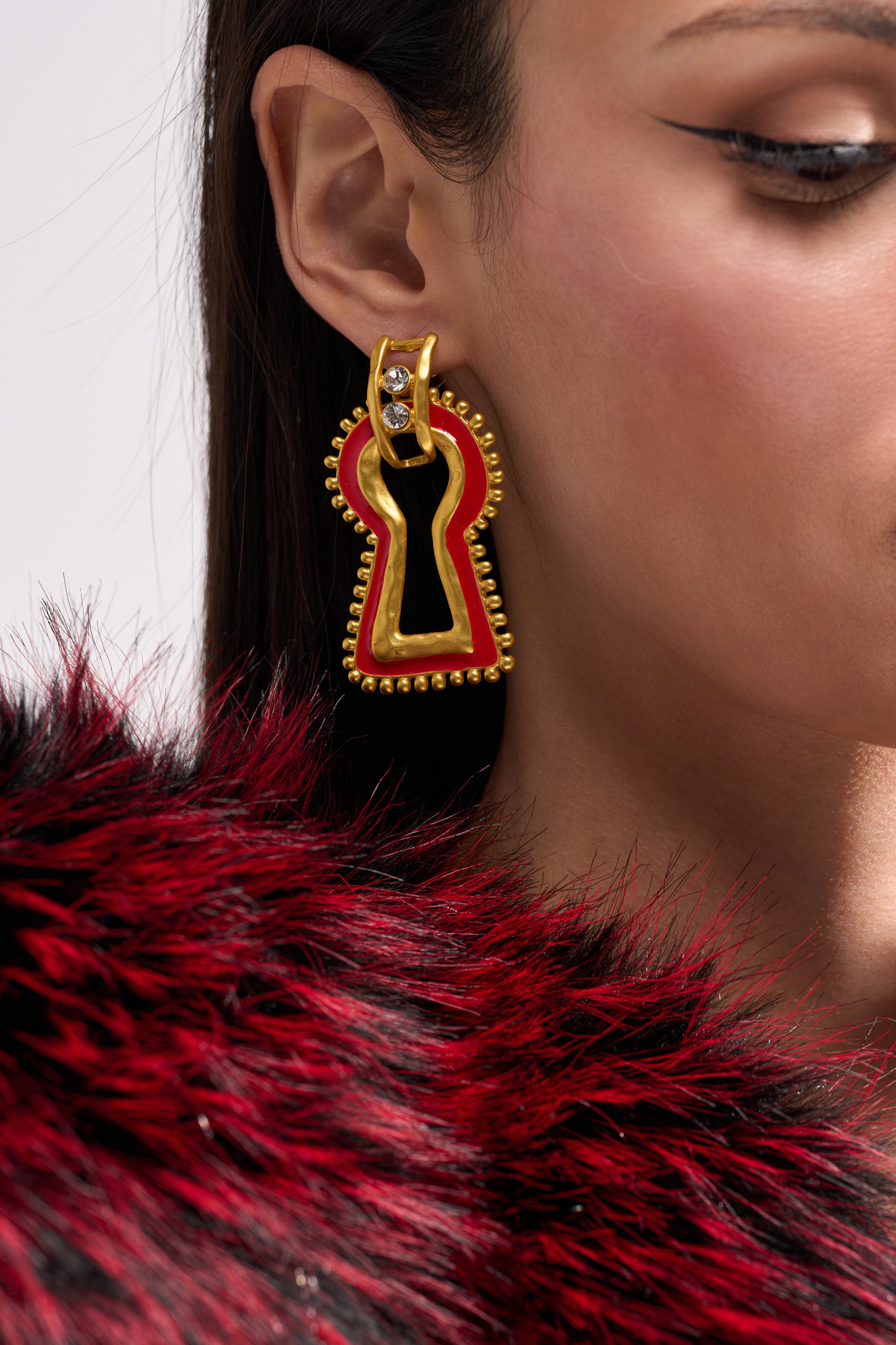 Close-up of a woman wearing the UNLOCK ME EARRING, a bold keyhole design with gold metallic accents and a clear gem. She has straight brown hair, winged eyeliner, and is styled in a red and black faux fur—ideal for statement earring lovers. 0