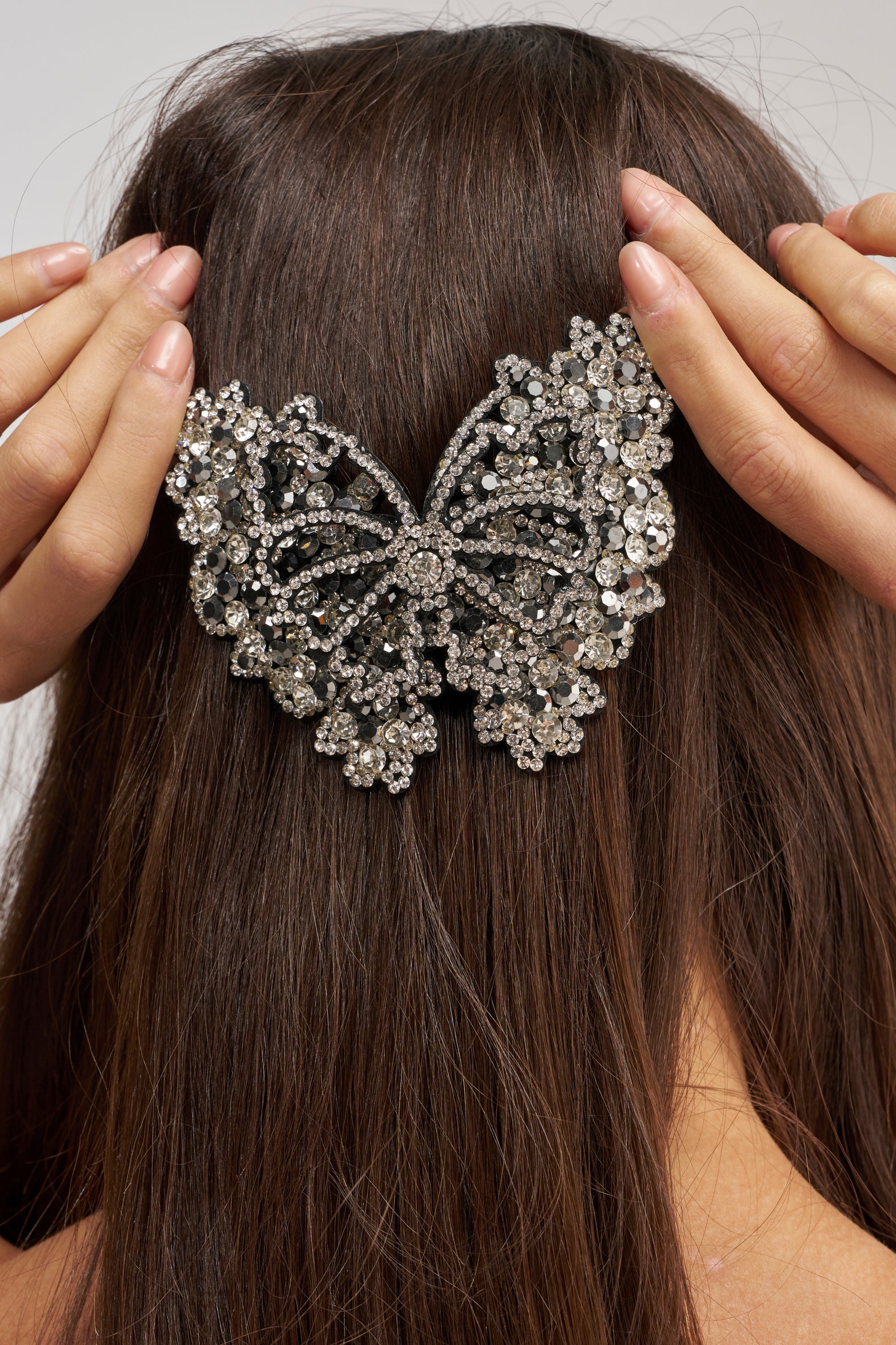 A person holds the FLY AWAY BUTTERFLY HAIR CLIP, a large rhinestone-studded hair accessory, against the back of their straight brown hair.