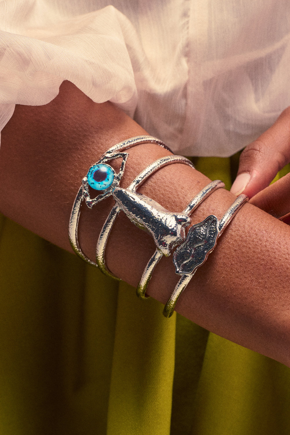The FACE IT BRACELET CUFF, a silver cuff bracelet featuring ocean-inspired designs and a blue stone in a boho-chic style, is worn on a person's forearm. The person pairs it with a white blouse and green skirt.