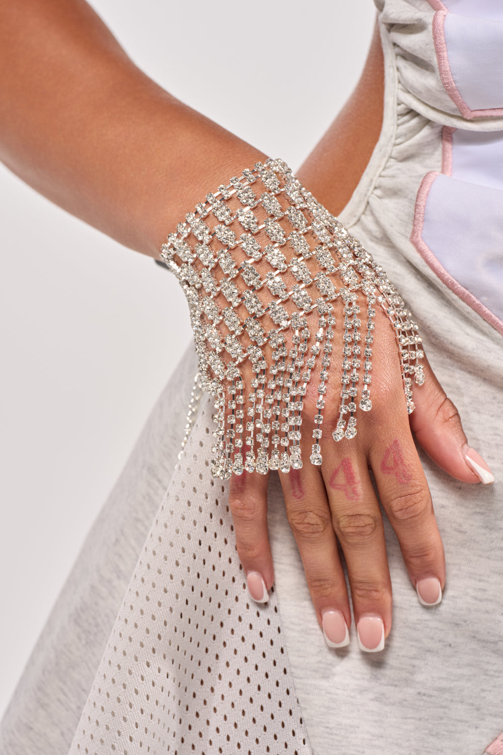 A close-up of someone wearing the TO THE PARTY BRACELET with sparkly rhinestone fringe and silver mesh. Their nails are painted pale pink, red marker numbers appear on their fingers, and they wear a light grey and white outfit.