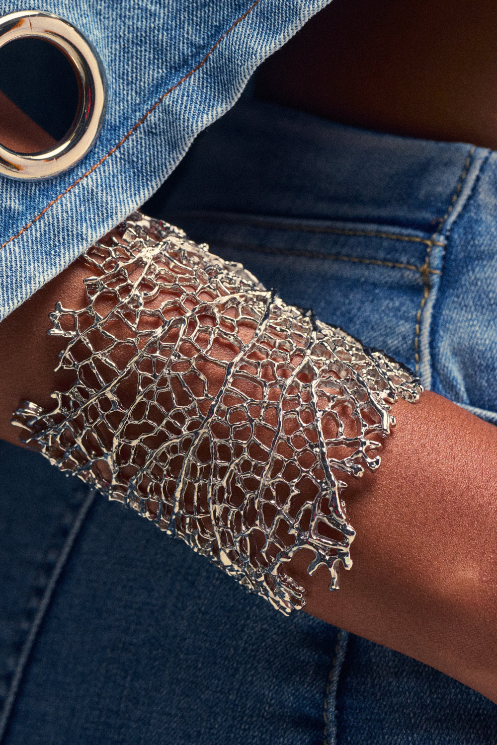 A close-up of the AT THE EVENT BRACELET CUFF IN SILVER, an intricately woven silver cuff bracelet worn on a person’s wrist and paired with denim clothing—this striking accessory is the perfect statement piece for summer.
