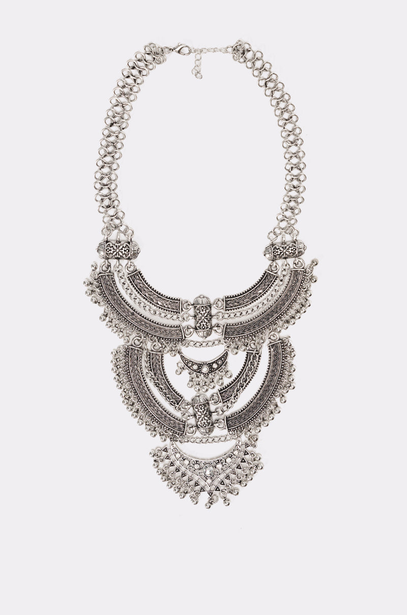 The AT THE FESTIVAL NECKLACE features three large, intricately patterned crescent-shaped silver tiers with engraved details and dangling beads, shown against a plain white background. 2