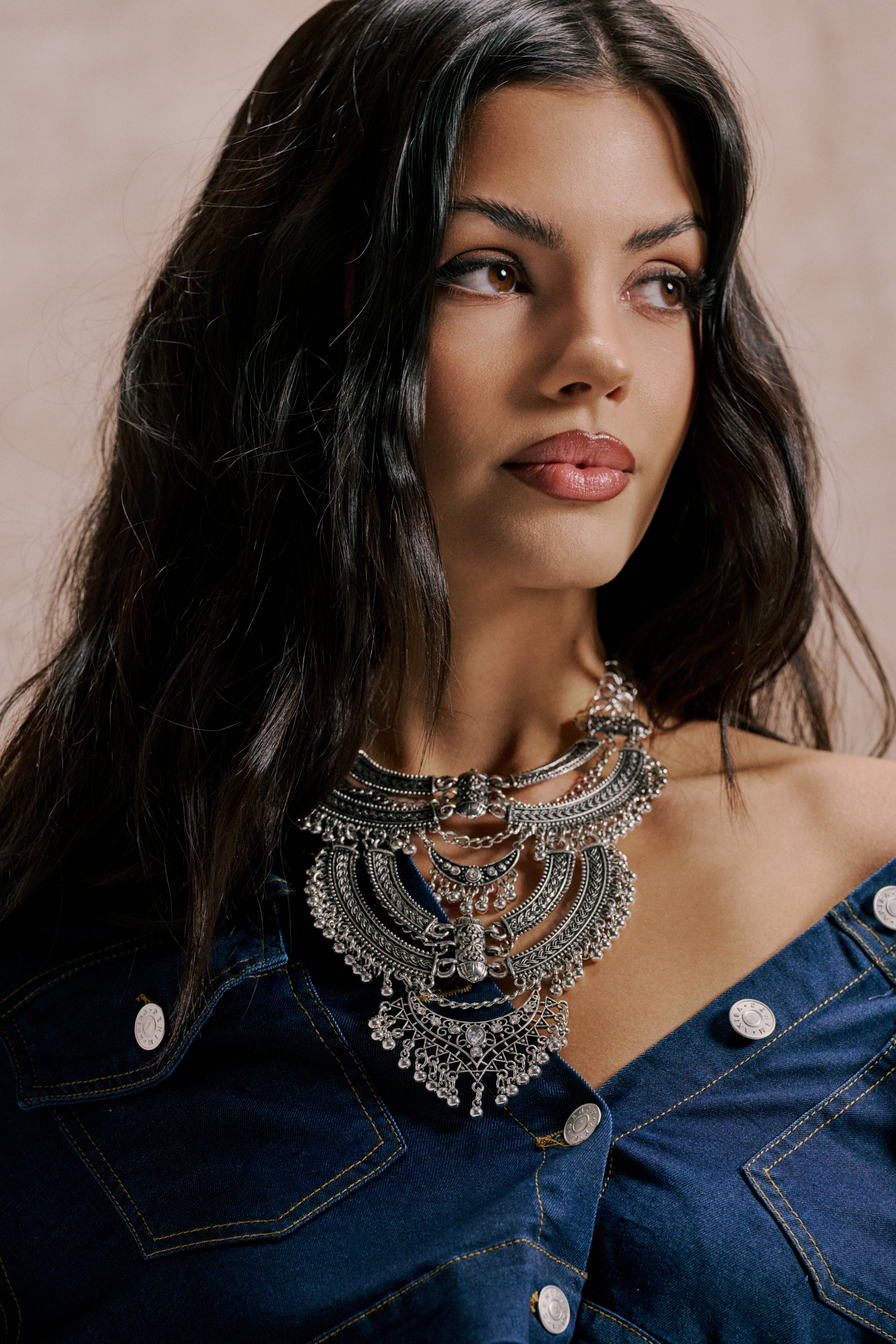 A woman with long dark hair wears the AT THE FESTIVAL NECKLACE, a large silver bib style, with a blue denim off-shoulder top. She looks to the side with a neutral expression against a plain background. 1