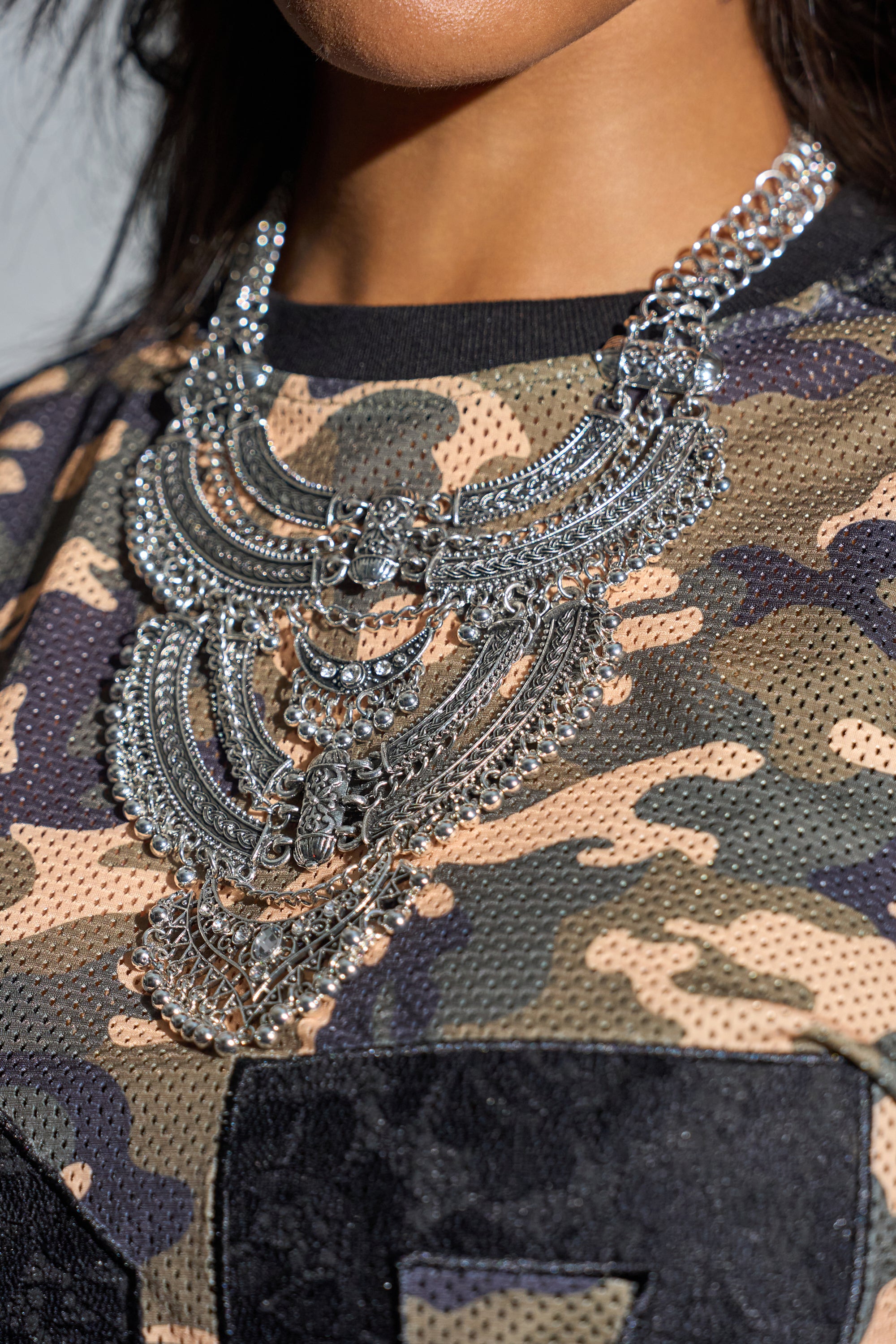 A person wears the AT THE FESTIVAL NECKLACE, layered over a camouflage mesh shirt. The cropped photo highlights the necklace’s bold, ornate design by focusing on the lower face, neck, and upper chest. 0