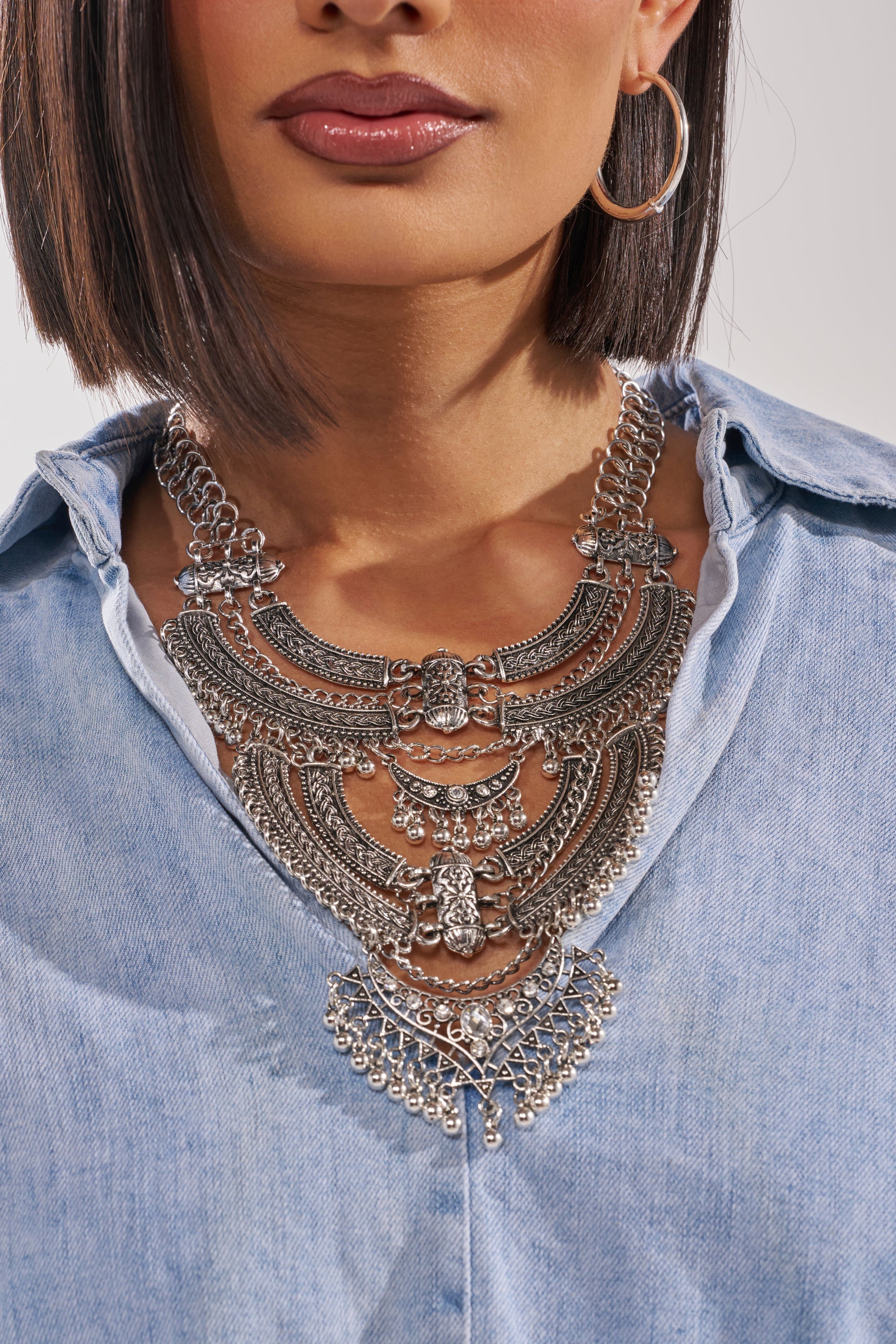 A woman models the AT THE FESTIVAL NECKLACE—a bold, boho-inspired silver bib with intricate designs and dangling details—paired with a light denim shirt and large silver hoop earrings. Only her lower face and upper torso are shown. 3