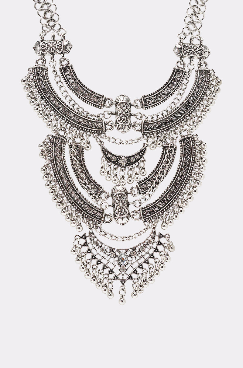 The AT THE FESTIVAL NECKLACE is a detailed silver bib necklace with three crescent-shaped layers, ornate designs, and dangling beads, shown against a white background. 5