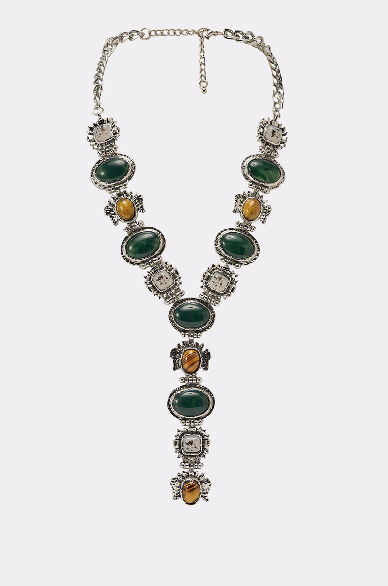 The IM OUTSIDE NECKLACE is a vintage silver statement piece with ornate settings, featuring oval green stones, square clear stones, and brown-yellow stones in a Y-drop arrangement on a white background. 0
