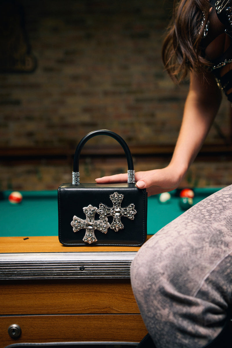 A person sits on the edge of a pool table, resting one hand near the EVERYTHING CHROME PATCH PURSE, which is adorned with rhinestones and jeweled crosses. Pool balls are scattered on the table against a brick wall background.
