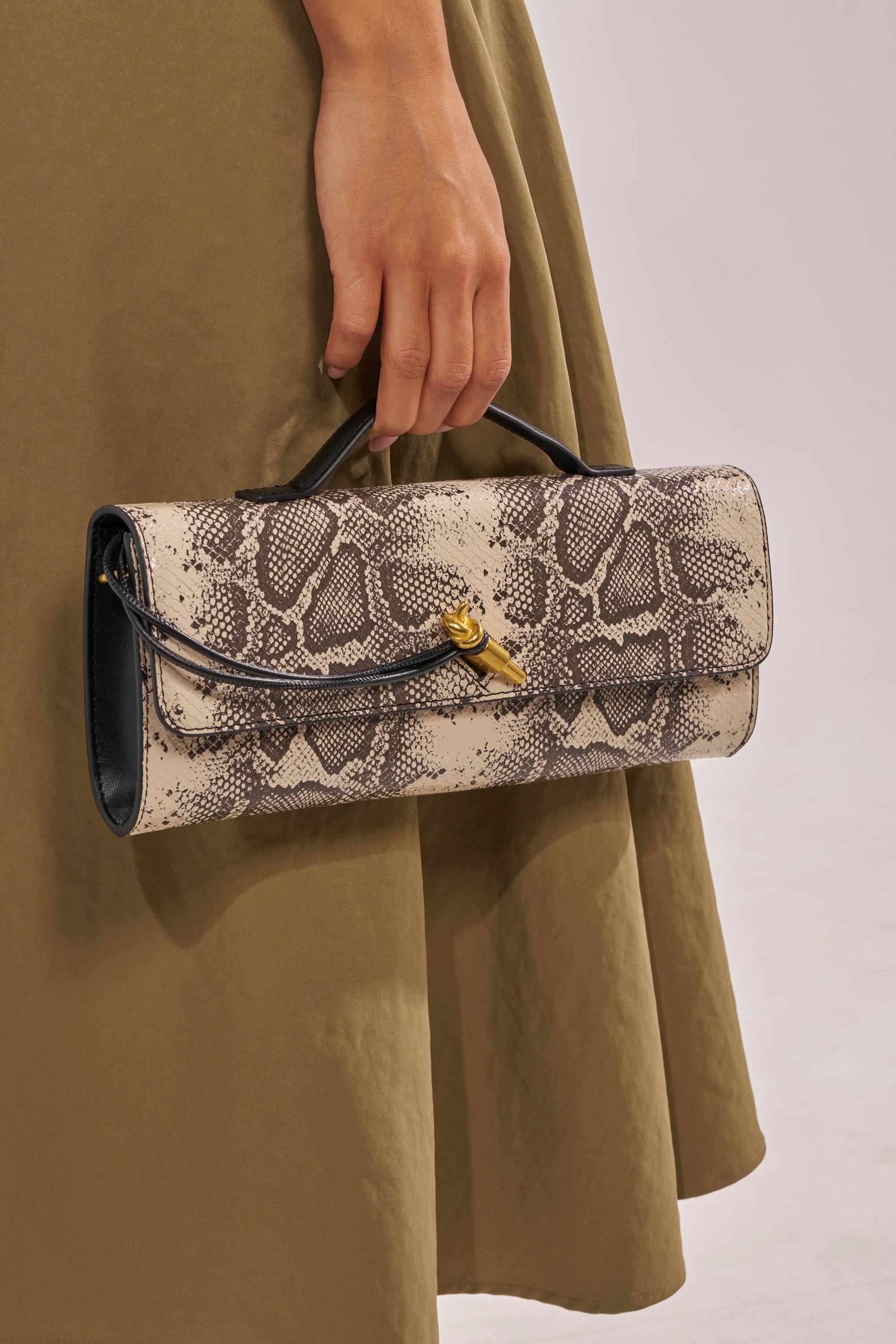 A person in an olive green skirt holds the I BITE PURSE—a chic clutch with a black and beige snakeskin print, short handle, and gold clasp. 1