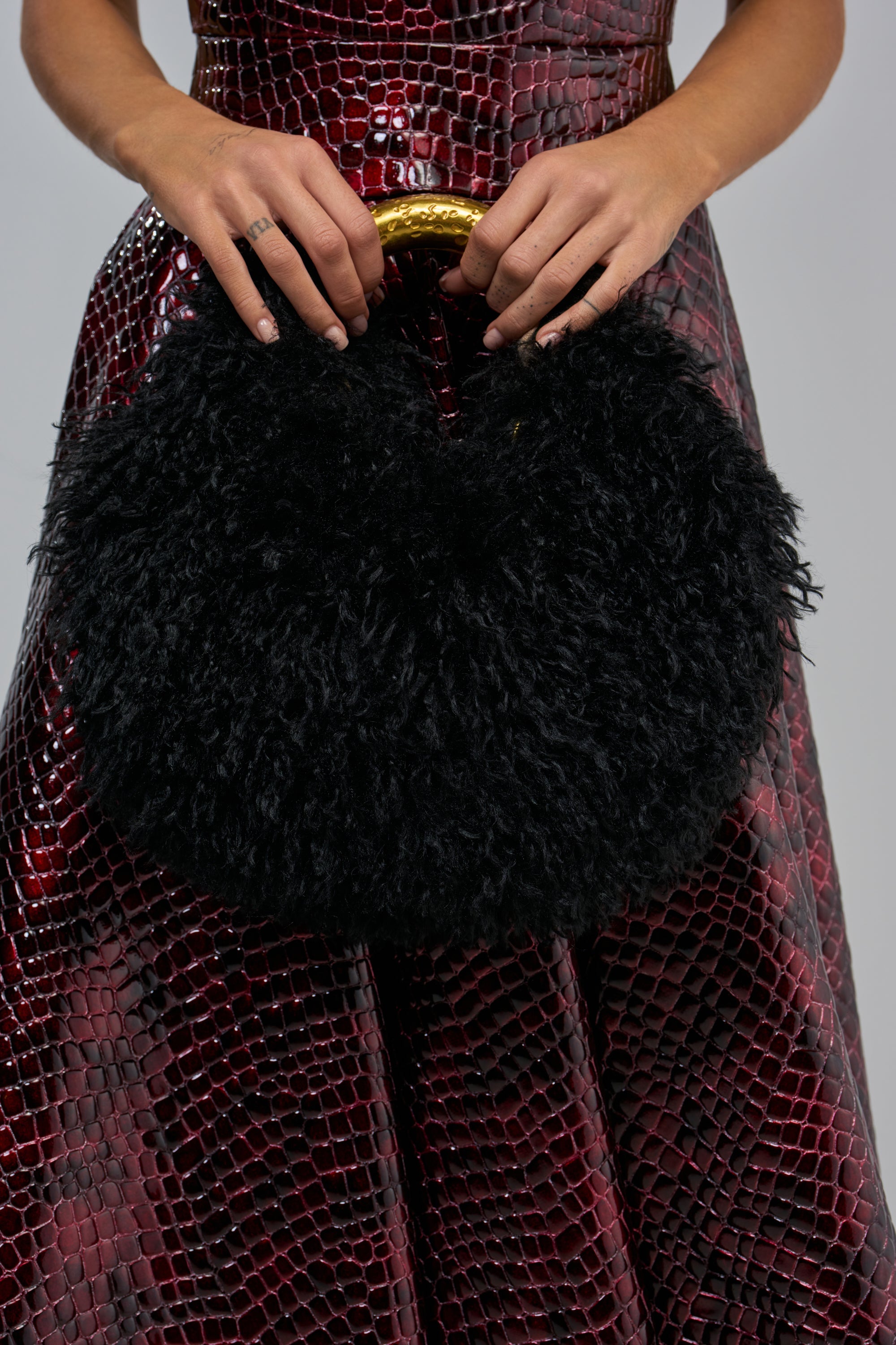 A person holds a SO FURRY BAG, a crescent-shaped, black faux-fur handbag with a metallic gold handle, in front of a deep red, crocodile-textured dress.