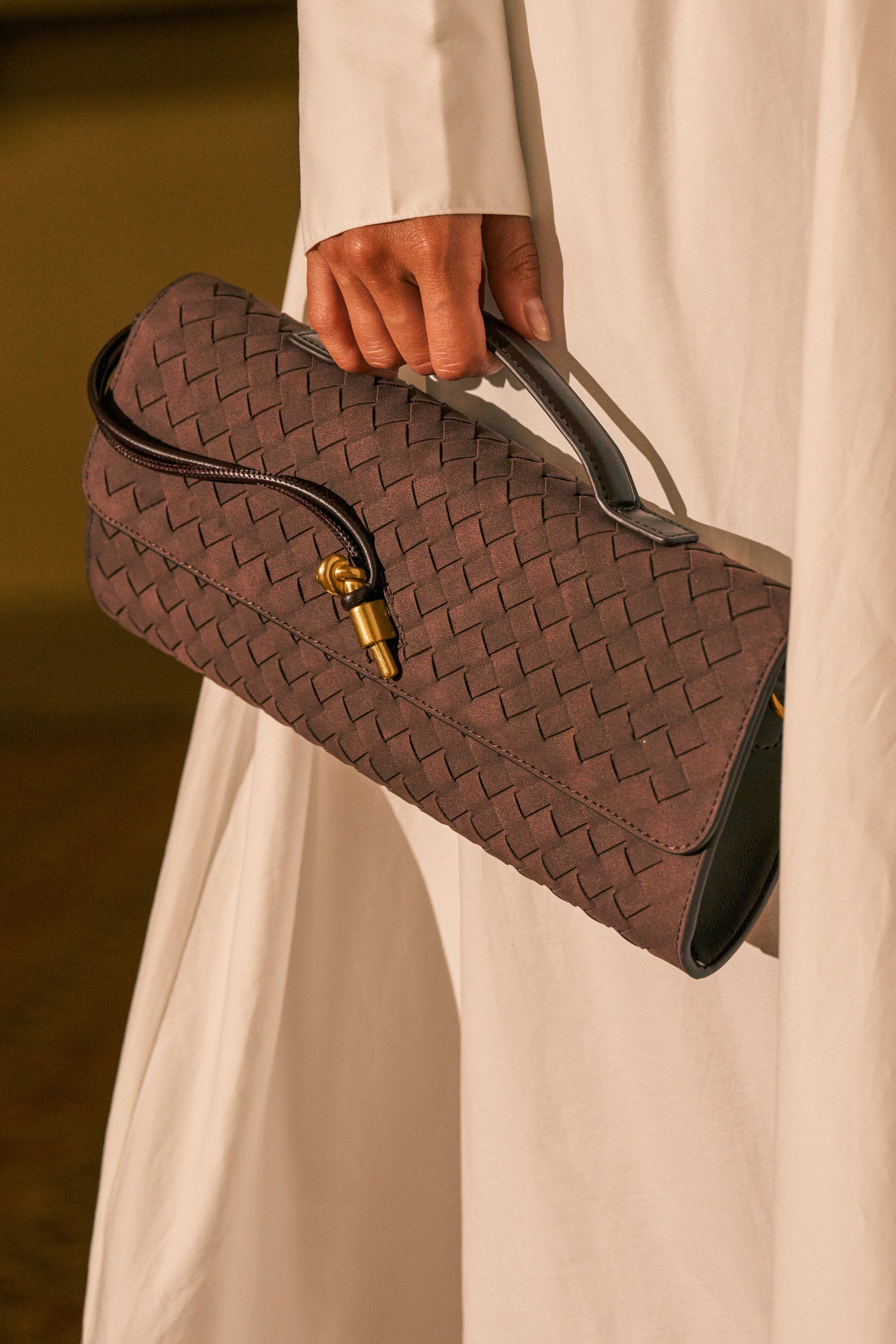 A person in a white garment holds the TRY ME SUEDE BAG, which features a brown woven texture, a short handle, and a gold clasp.