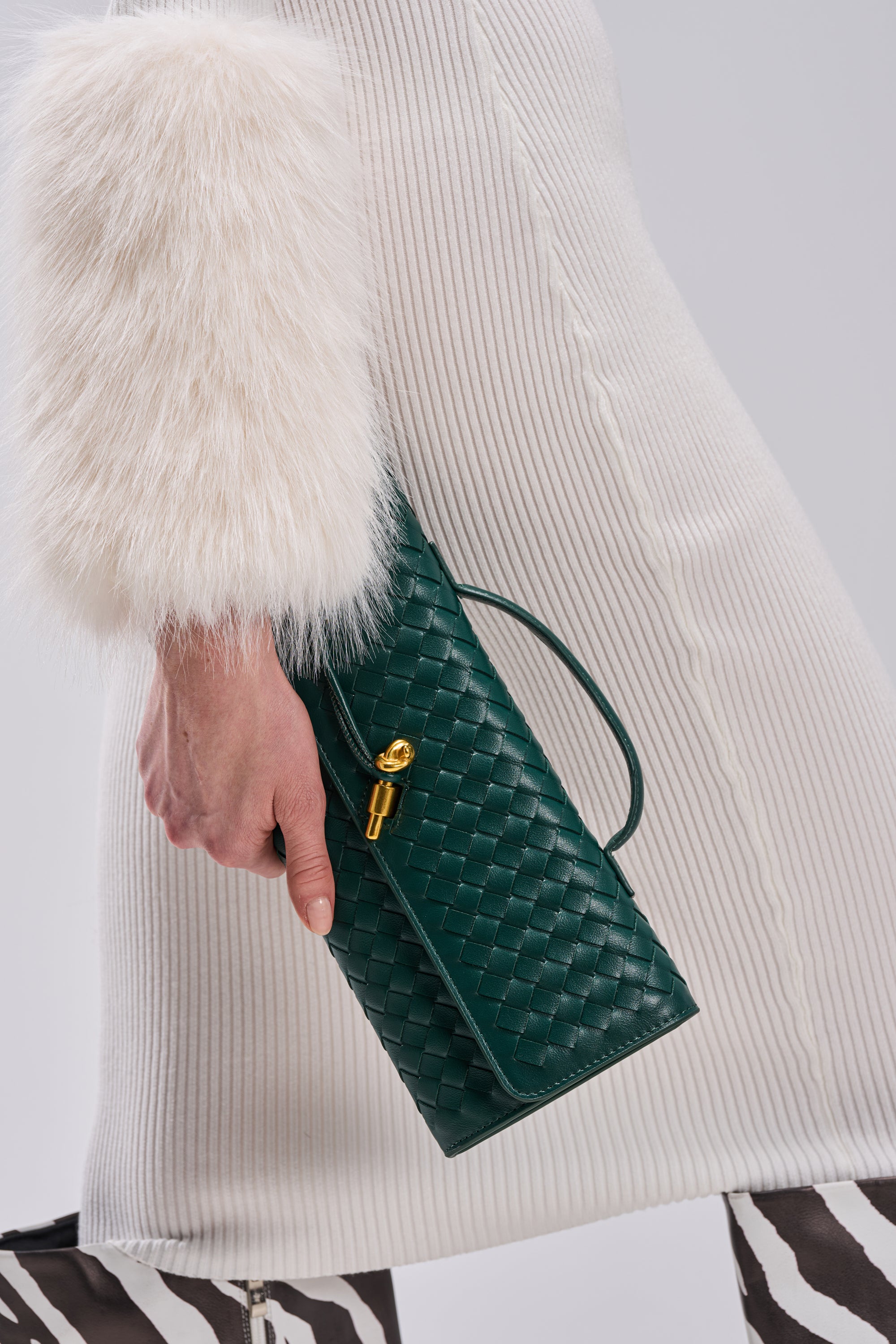 A person in a white textured coat with faux fur cuffs holds the LUXE LIFE PURSE, a dark green woven clutch featuring a gold clasp and crossbody strap. Zebra-striped pants complete the chic ensemble.
