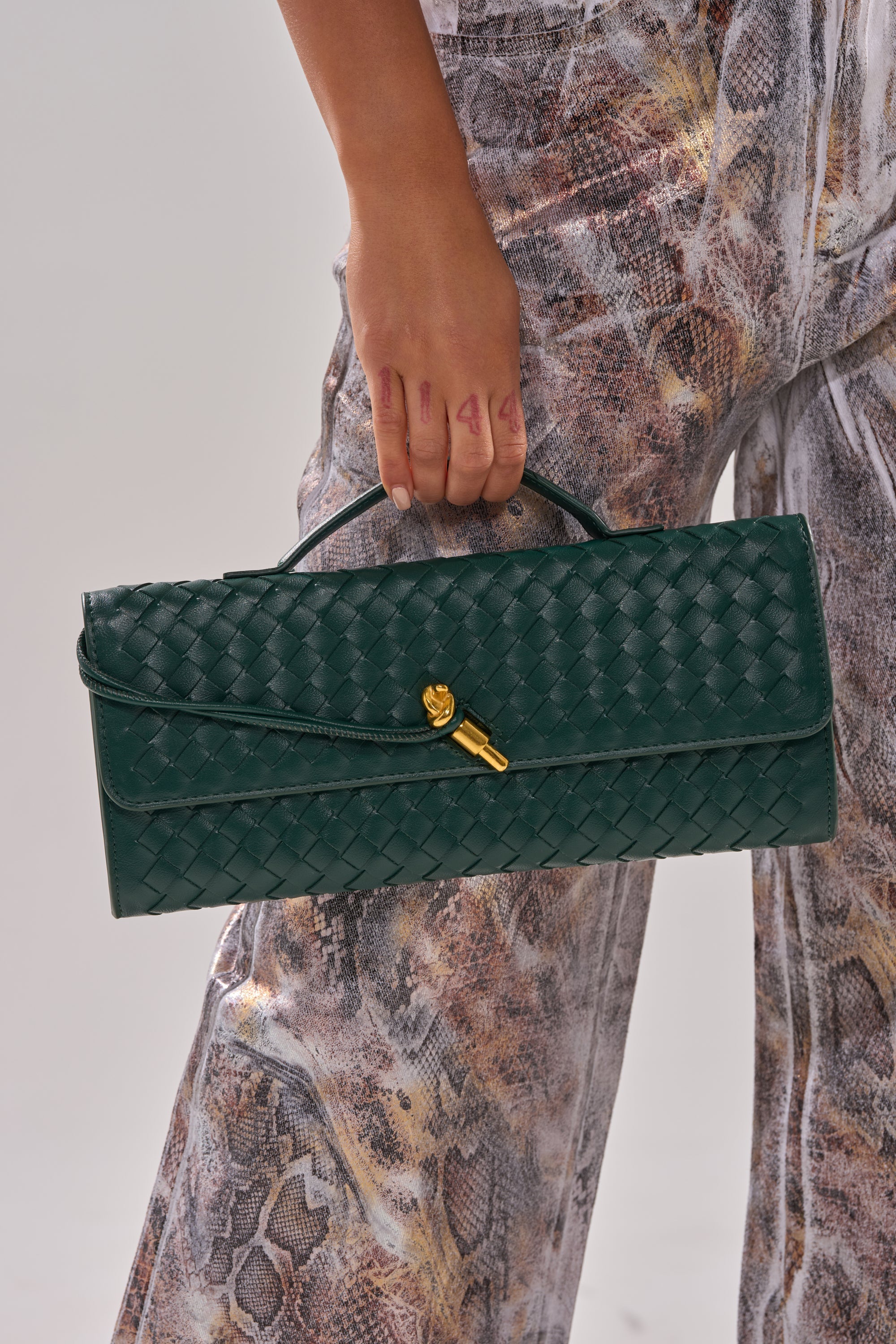 A person in snakeskin-patterned pants holds the LUXE LIFE PURSE—a dark green woven clutch with a gold clasp and handle, ideal for fans of chic faux leather purses. 2