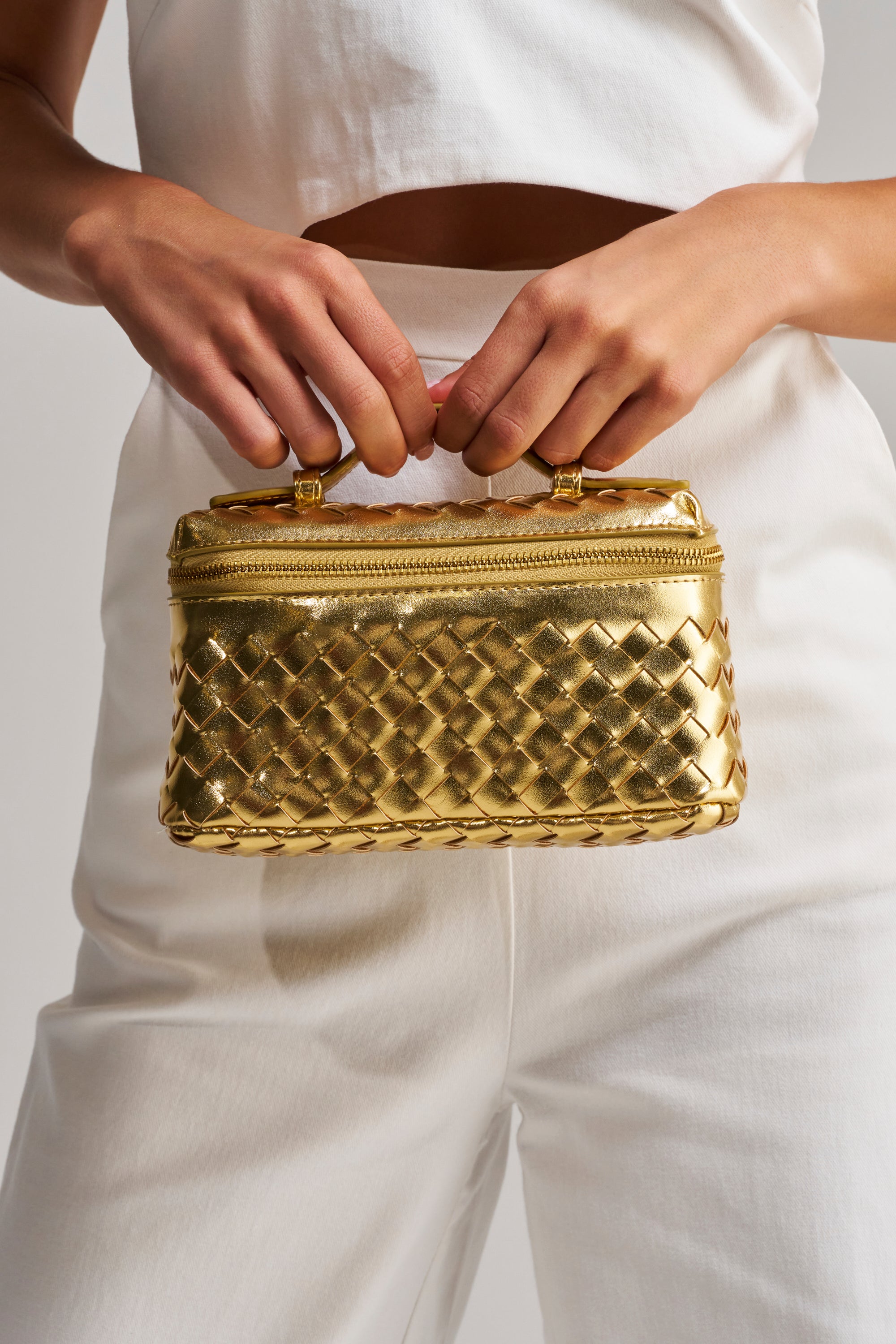 A person in white holds the GOLD ME CLOSER PURSE—a small, shiny gold metallic bag with a woven texture and zipper—perfect for transitioning from day to night.