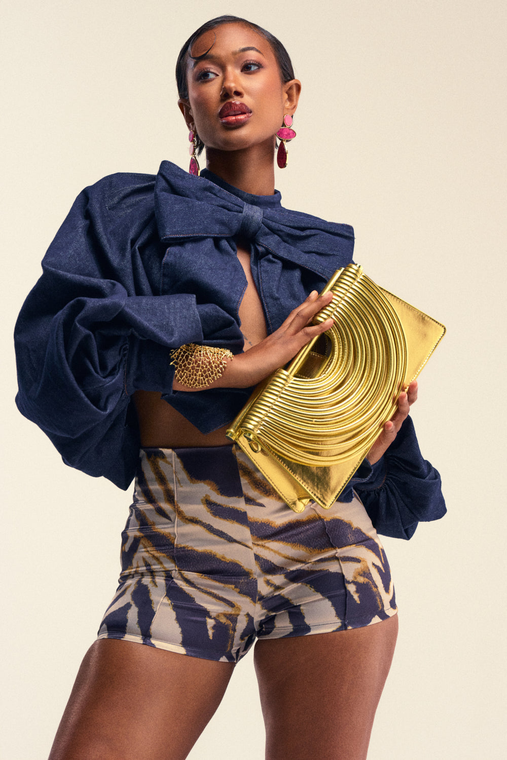 A woman poses confidently in a navy blouse with large bows, patterned shorts, pink earrings, a gold bracelet, and holds the GOLDEN HOUR PURSE against a neutral background.