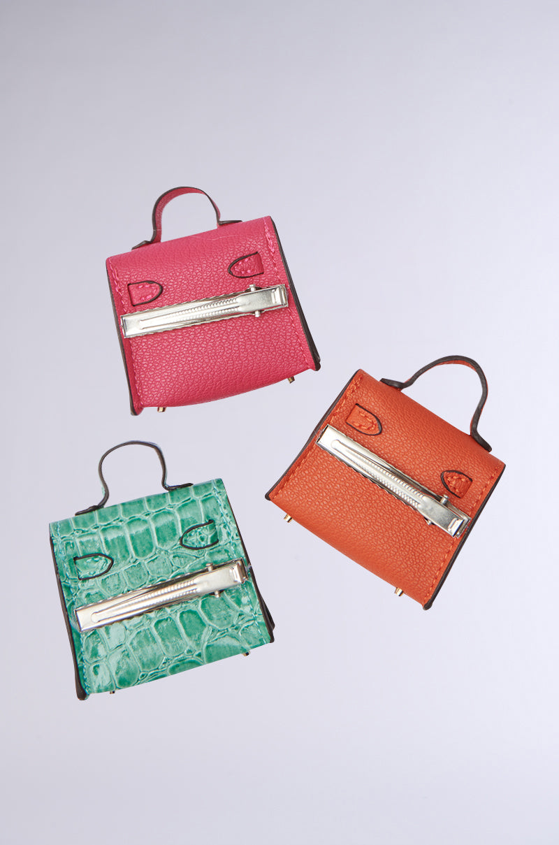 The STAY IN MY BAG HAIR CLIP SET showcases three faux leather small handbags in pink, orange, and green crocodile textures. Each bag has a metallic front zipper and a small top handle, arranged against a plain background.