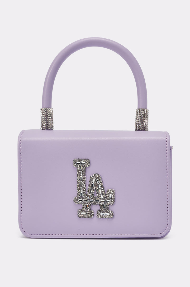 The IN THE CITY APPLIQUE PURSE is a small, rectangular lavender mini handbag with a sturdy handle and silver rhinestone "LA" logo on the front—chic, sparkly, and perfect for any occasion. Background is plain white. 0